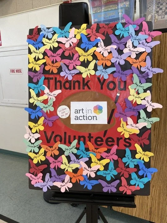 A red poster with the words 'Thank You Volunteers' in black, decorated with numerous colorful paper butterflies with handwritten names and messages.