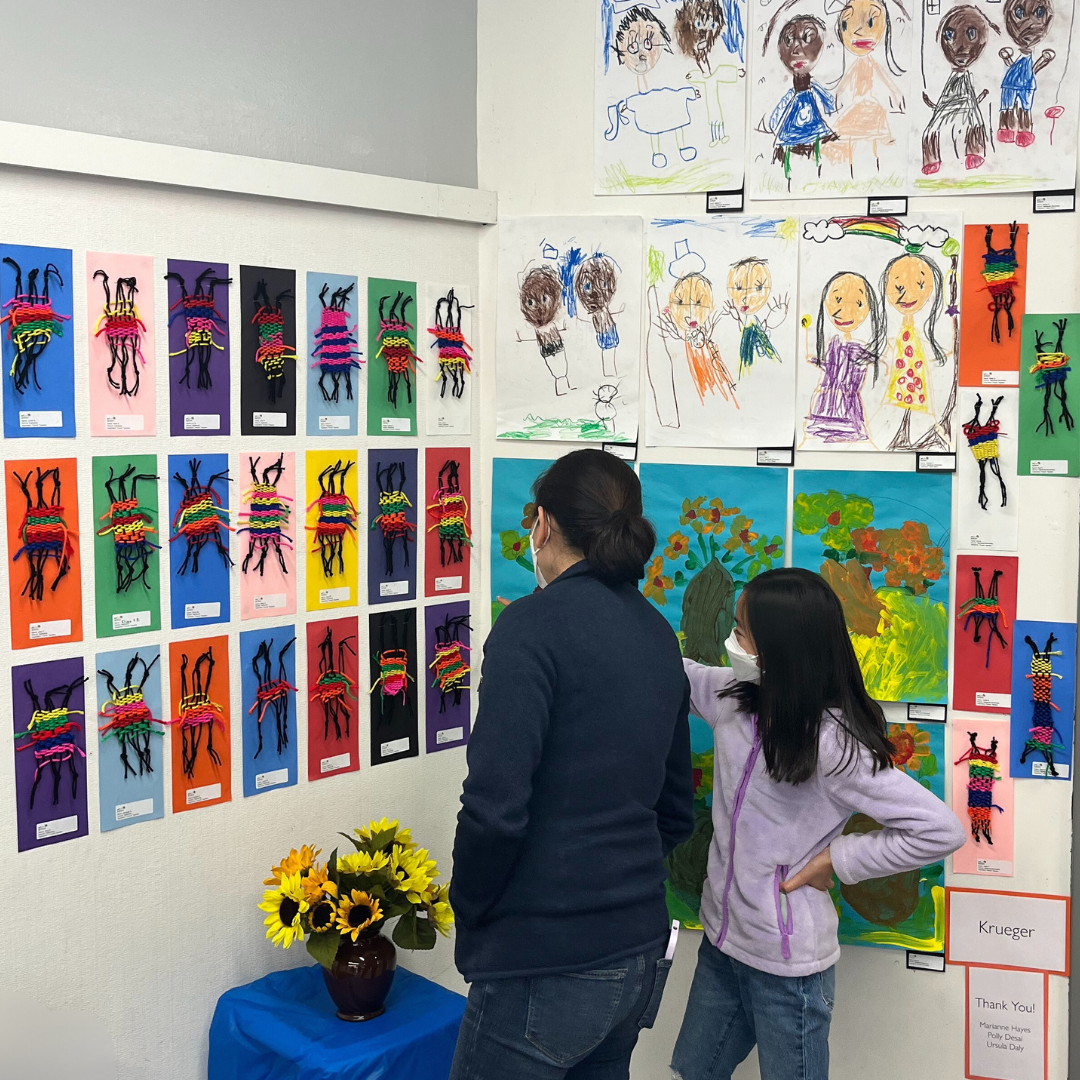 A woman and daughter wearing masks at an art exhibit, looking at colorful artwork and photographs of children's drawings on the wall.