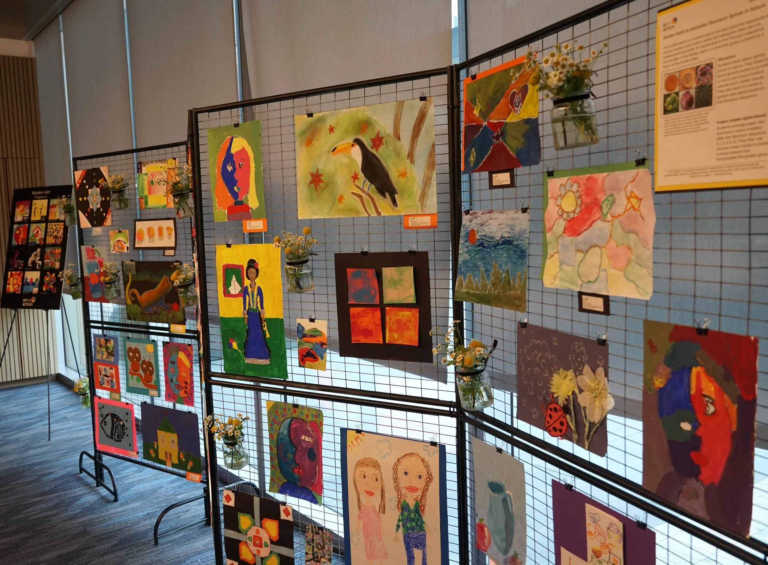 An art exhibit showcasing colorful children's paintings and drawings displayed on a black metal grid with small jars of flowers at the bottom, set in a brightly lit indoor space.