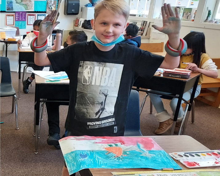 Photo of a 9 year old boy in a NBA shirt on. Both hands up with paint brush in the left hand and paint on hands. Painting on the desk below him.