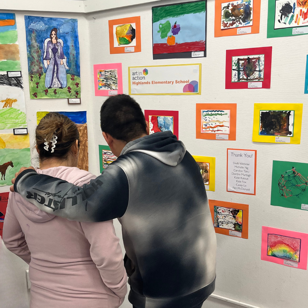 Two people viewing children's artwork at Highland Elementary School art exhibit, with colorful paintings and drawings displayed on the wall.