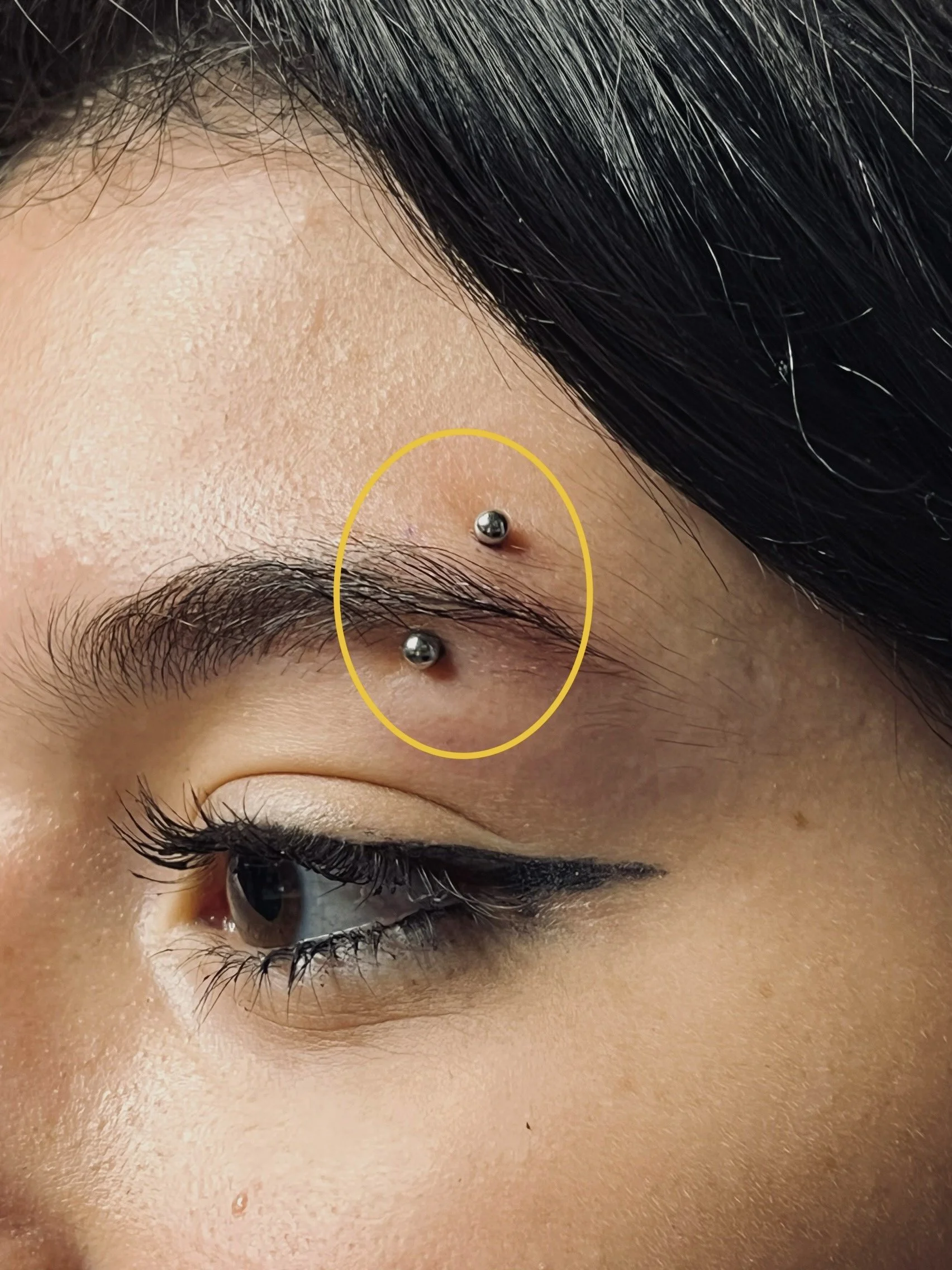 Eyebrow piercing on a person's face, with a metallic barbell and circular yellow highlight around the piercing area.