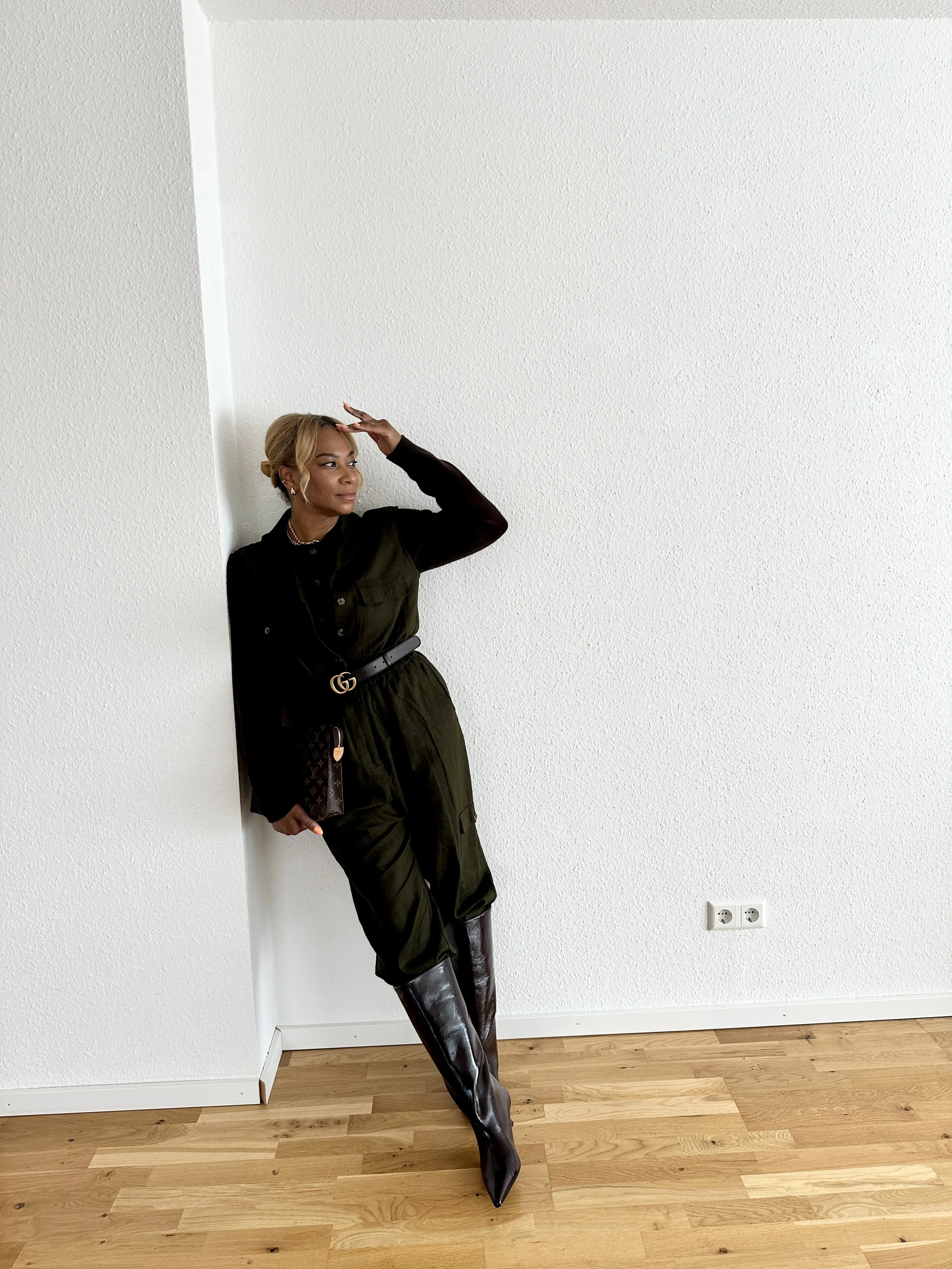 Young woman in black outfit and high black boots posing against a white textured wall with wooden floor and power outlet.