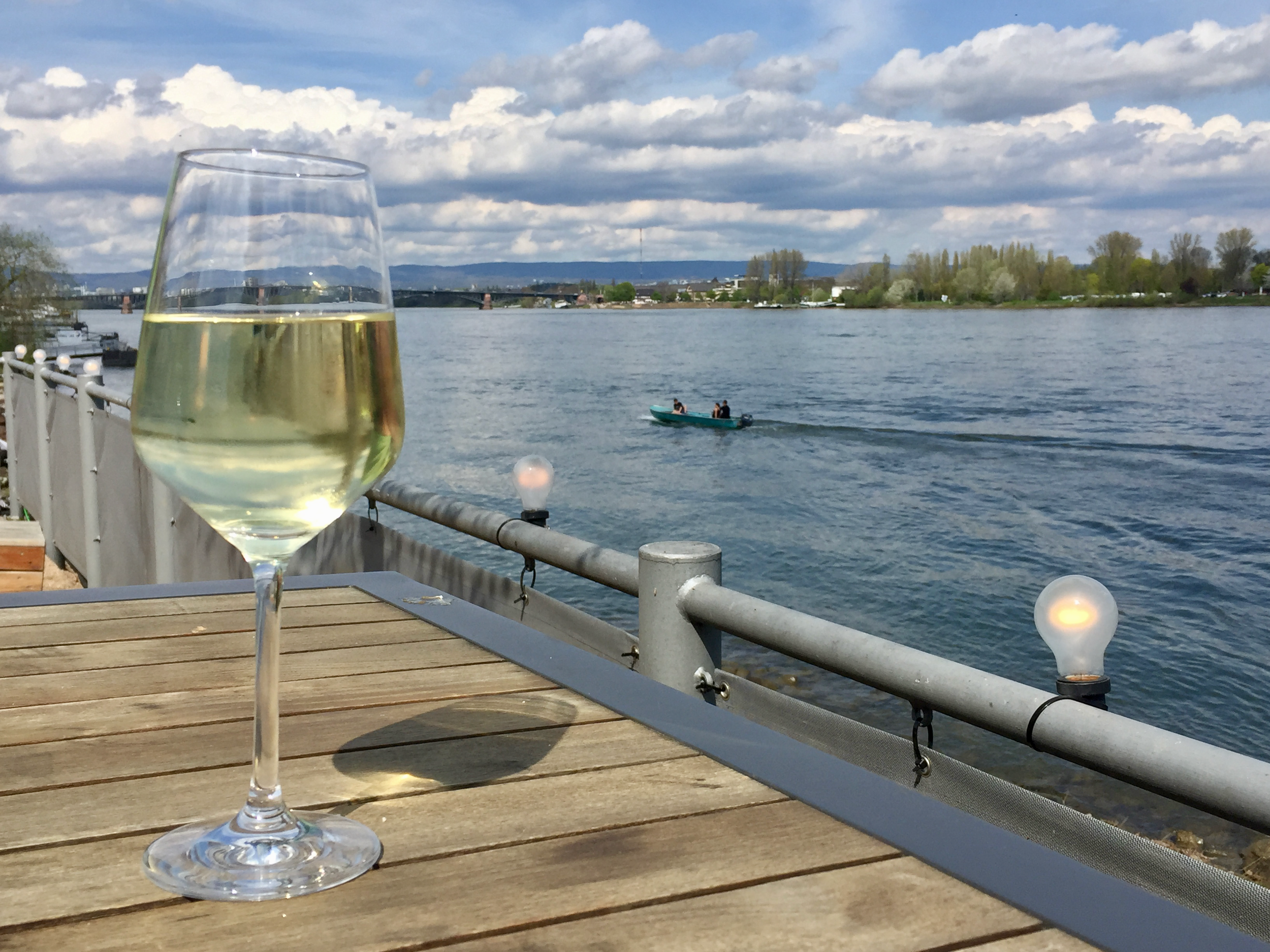 A glass of white wine on a wooden table by a river with boats and a bridge in the background, cloudy sky.