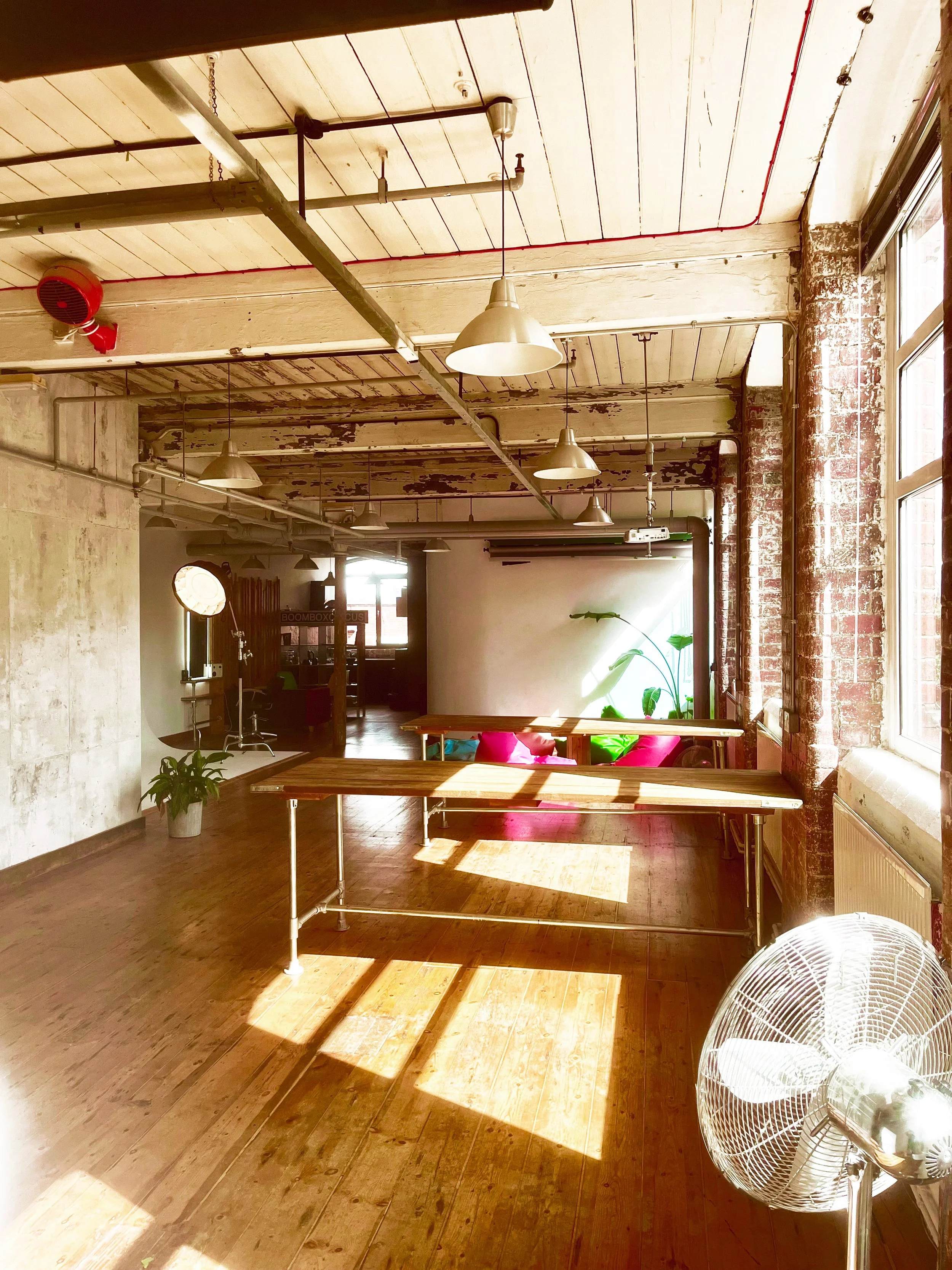 Bright industrial-style interior with wooden floors, exposed brick walls, large windows letting in sunlight, hanging ceiling lights, and seating area with pink and green cushions.