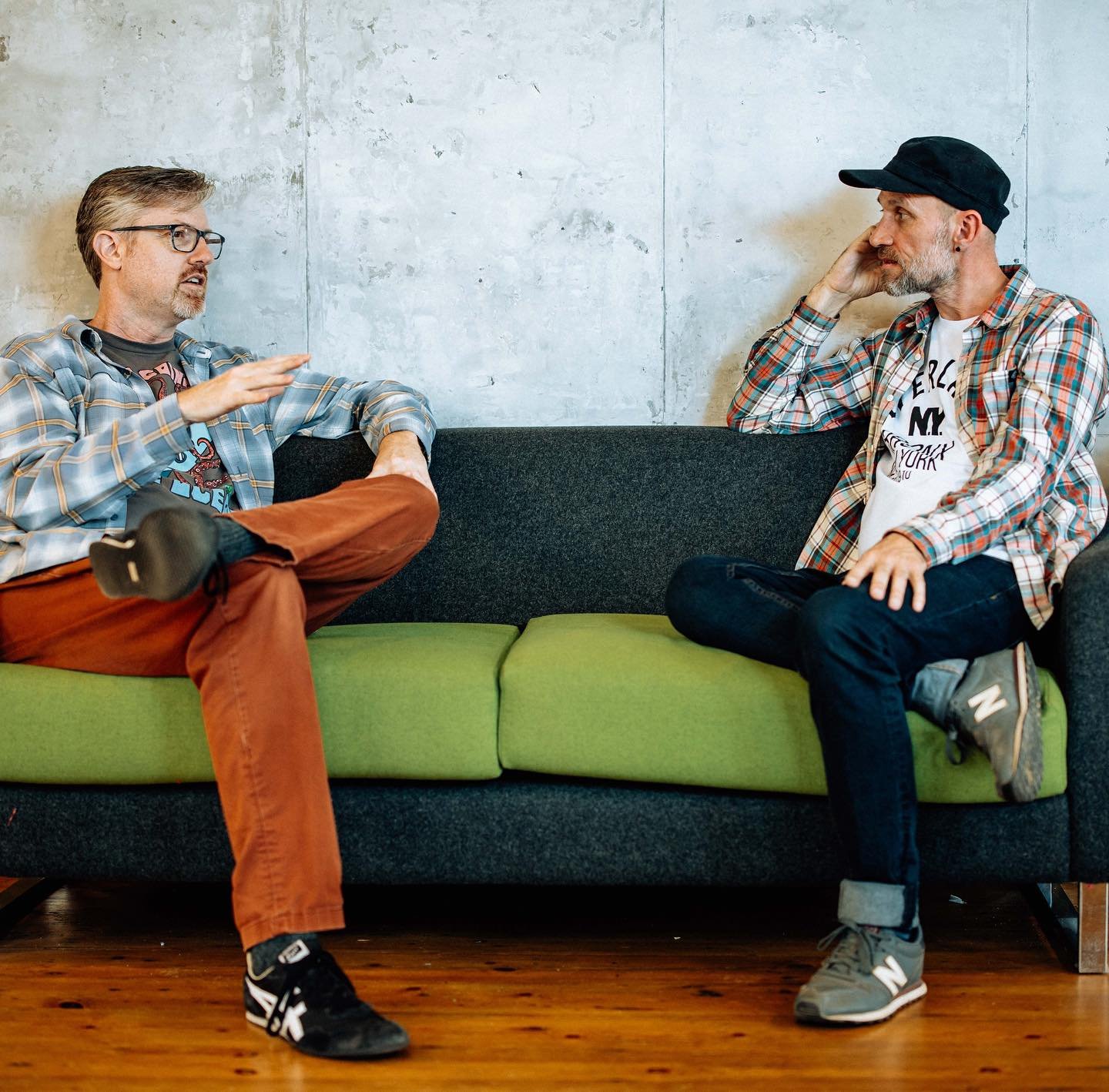 Two men having a conversation while seated on a green and black couch in a room with a plain wall. One man is gesturing as he speaks, the other listens with his hand on his head.