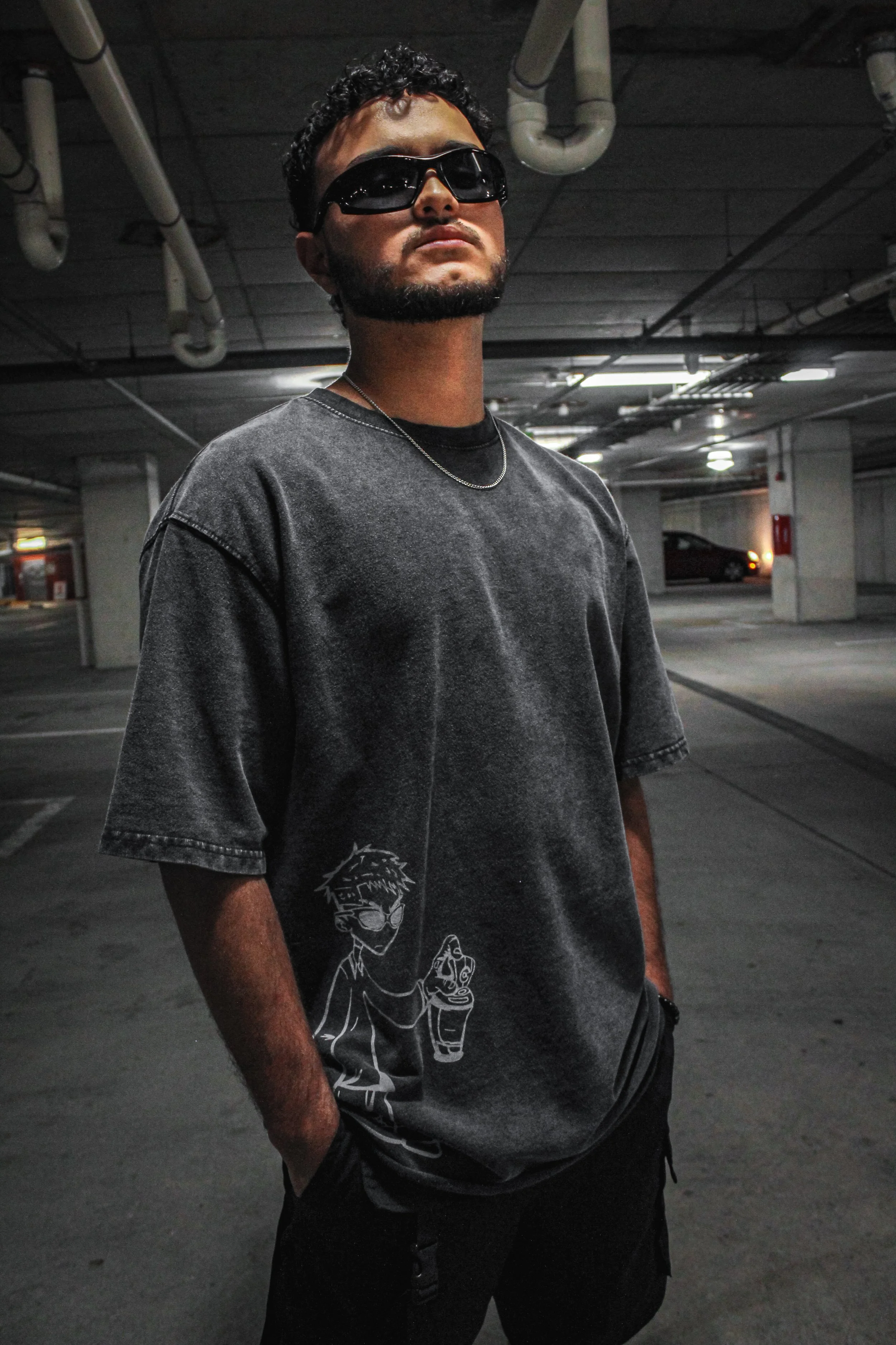 A young man standing in a parking garage, wearing black sunglasses, a dark gray T-shirt with a cartoon graphic on the bottom left, and black shorts, with hands in pockets.