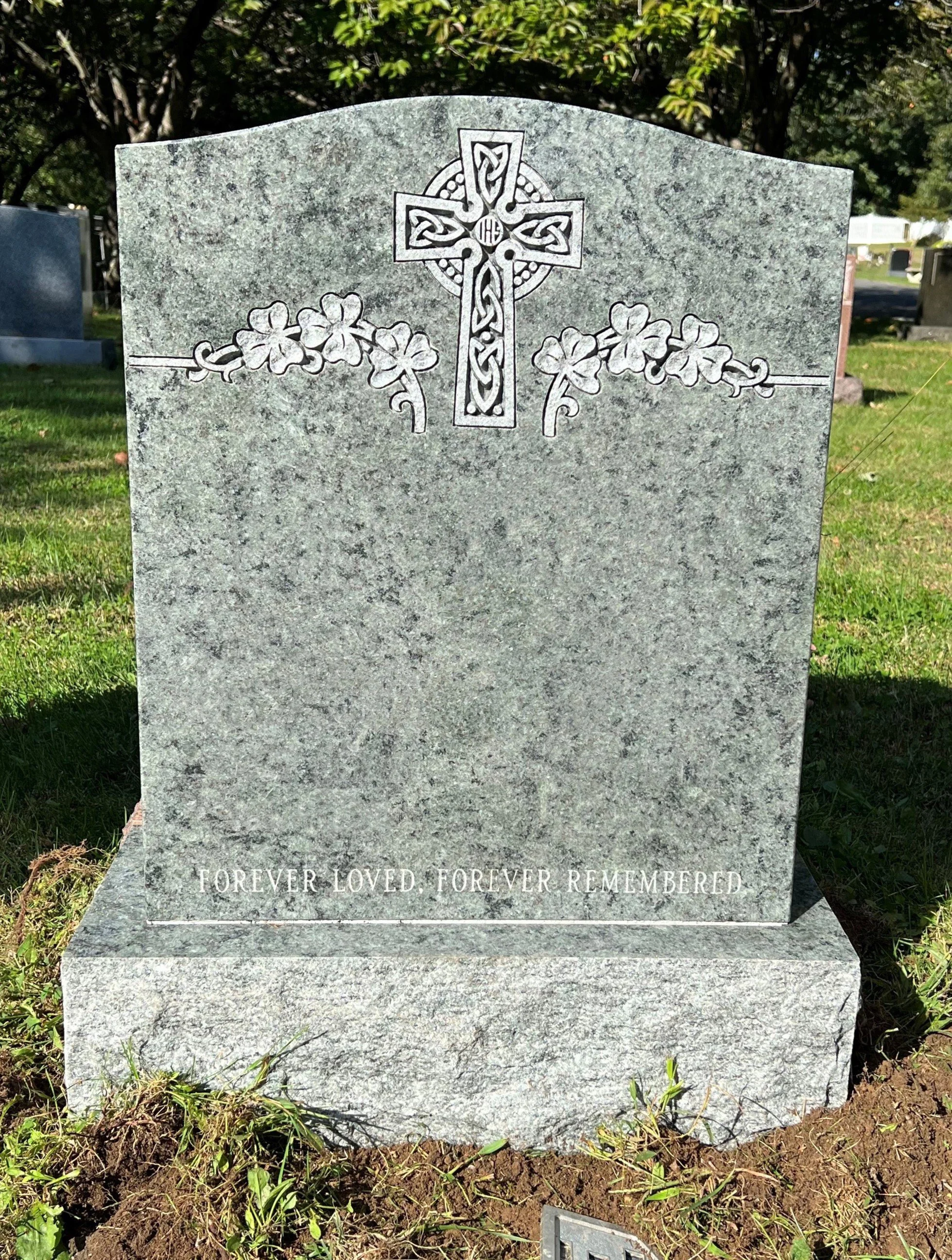 Gray granite headstone with a carved Celtic cross and shamrock designs at the top, and the inscription 'FOREVER LOVED. FOREVER REMEMBERED' at the bottom, in a cemetery with trees and other headstones in the background.