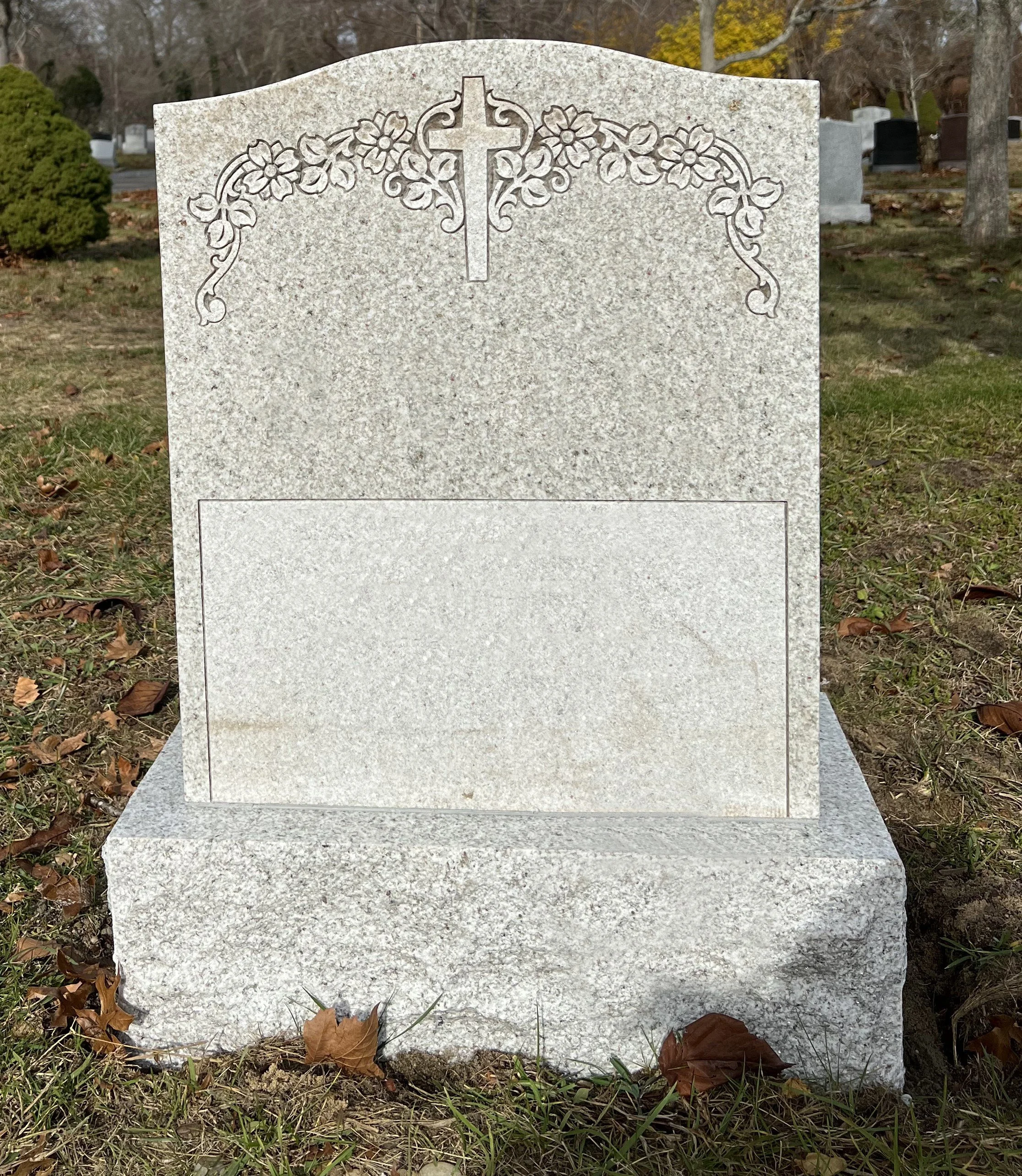 Gray granite headstone with engraved flowers and a cross at the top, located in a cemetery with fallen leaves and other headstones in the background.