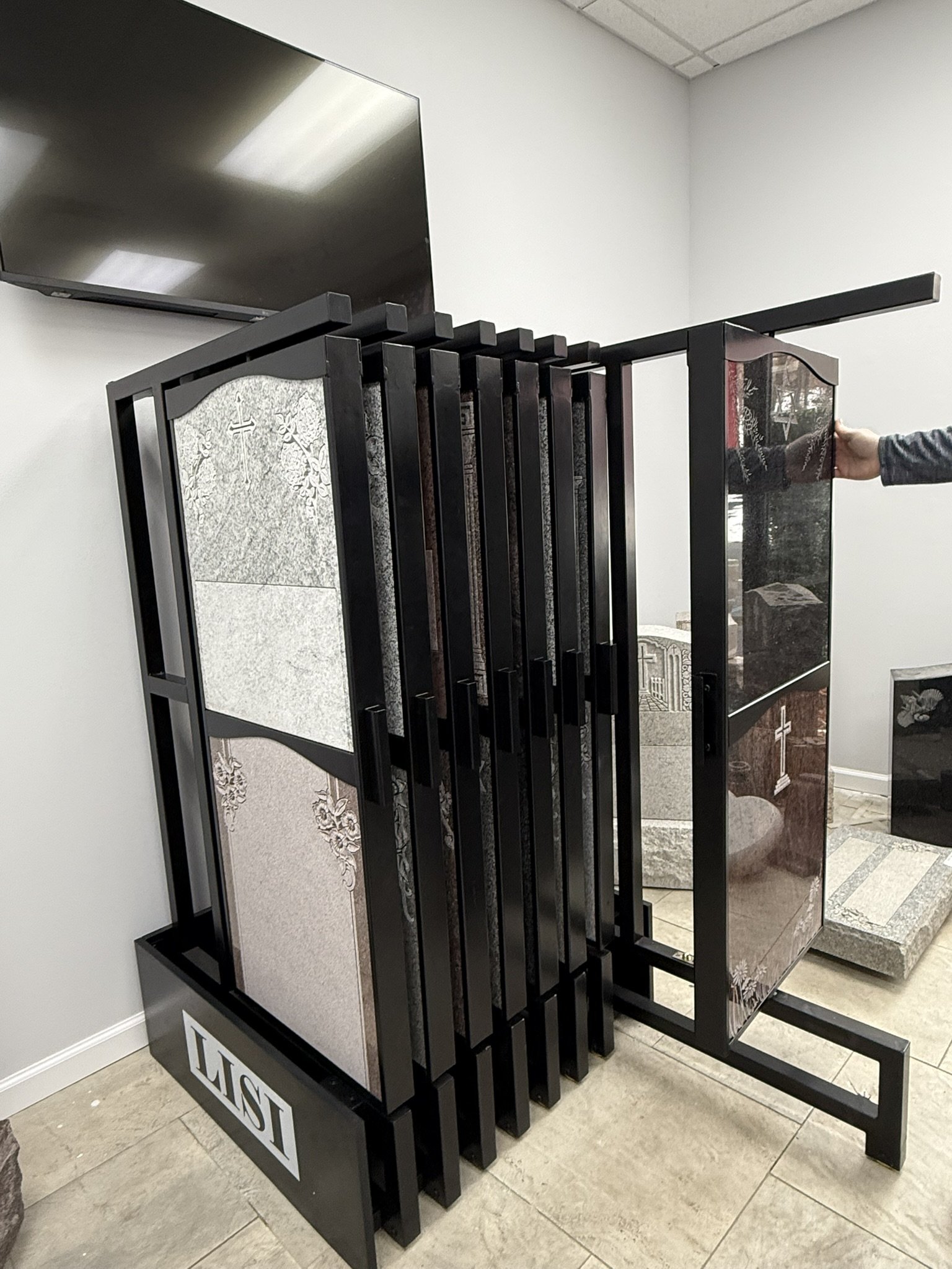 Black metal display rack with glass panels showing gravestones, with a flat-screen TV on the wall above.