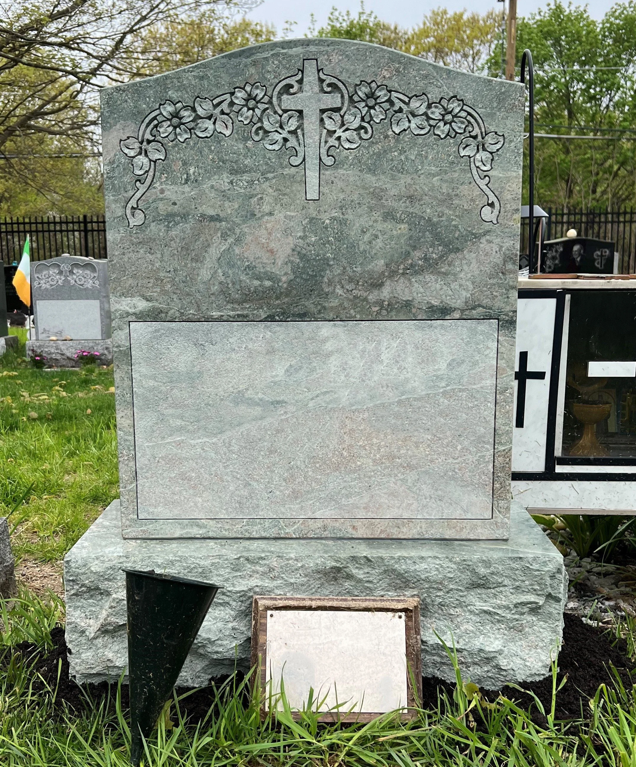 Gray marble headstone with floral and cross carvings at the top, a large blank rectangular plaque for inscriptions, and a smaller blank plaque at the base, located in a grassy cemetery.