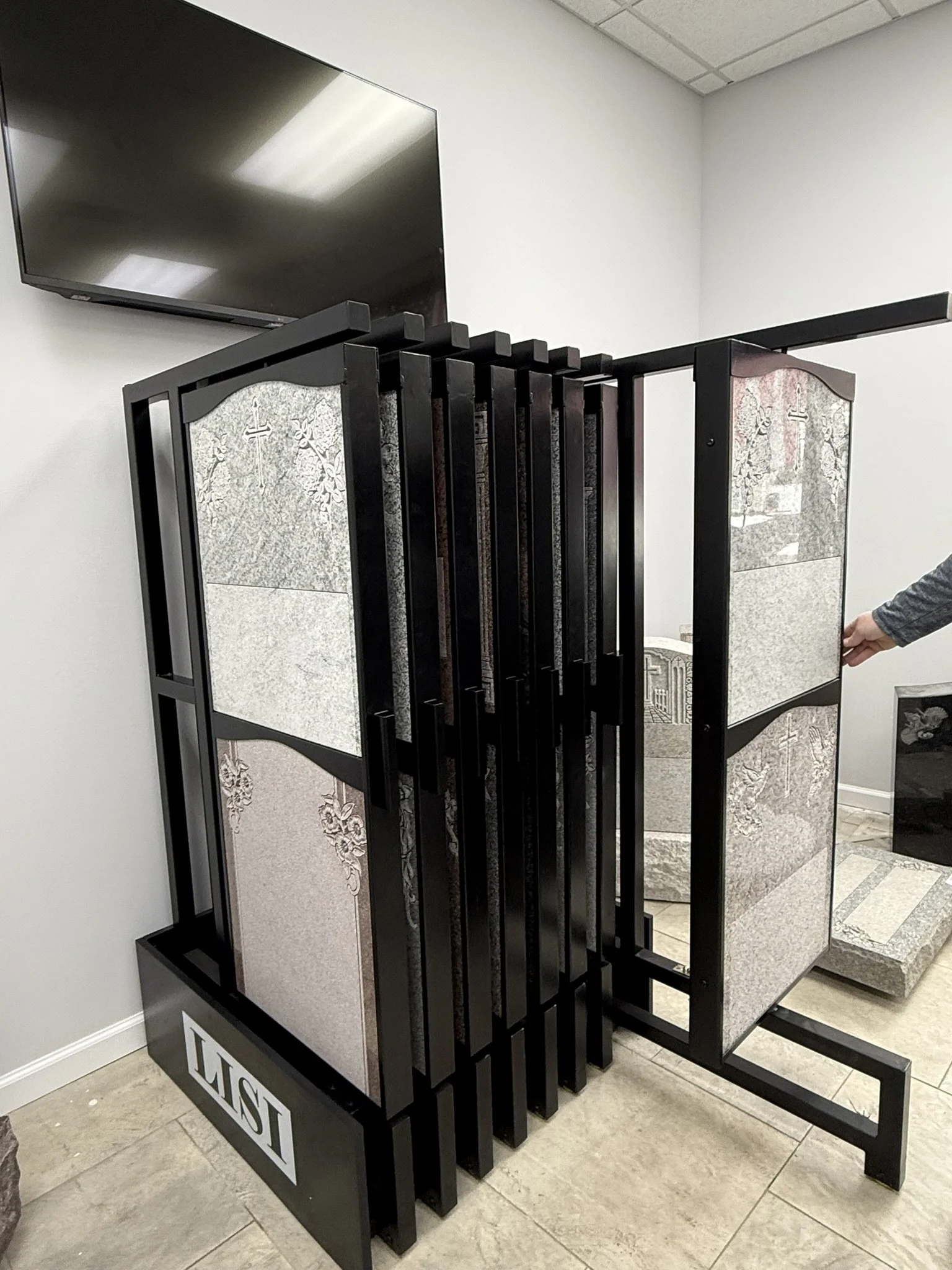 Display rack holding tombstone samples in a showroom, with a large flat-screen TV mounted on the wall above.