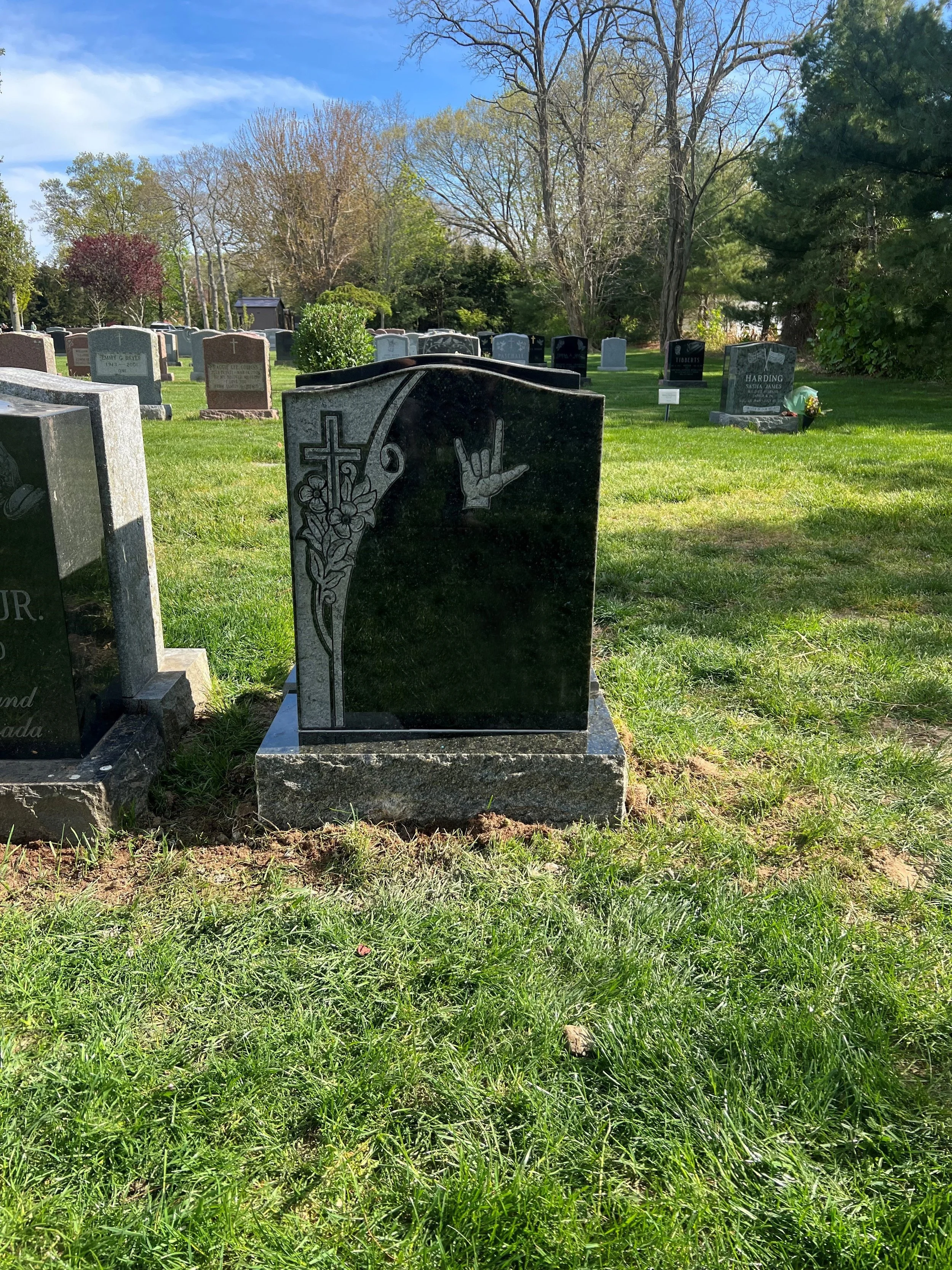 A black granite headstone in a cemetery, decorated with a cross and flowers engraving, featuring a carved hand making the sign of the horns.