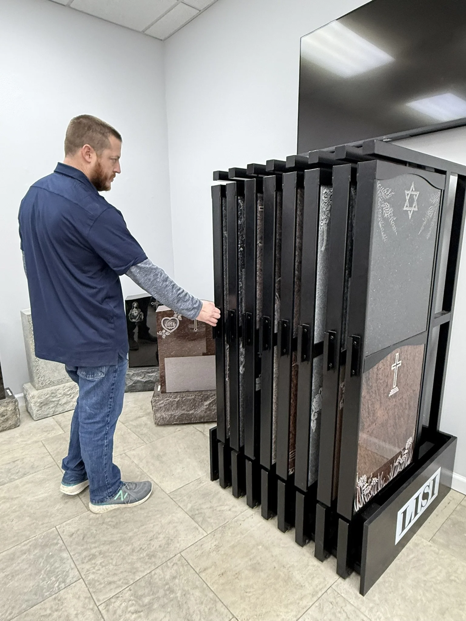Man in navy blue shirt looking at a display of multiple granite headstones in various colors and designs in a showroom.