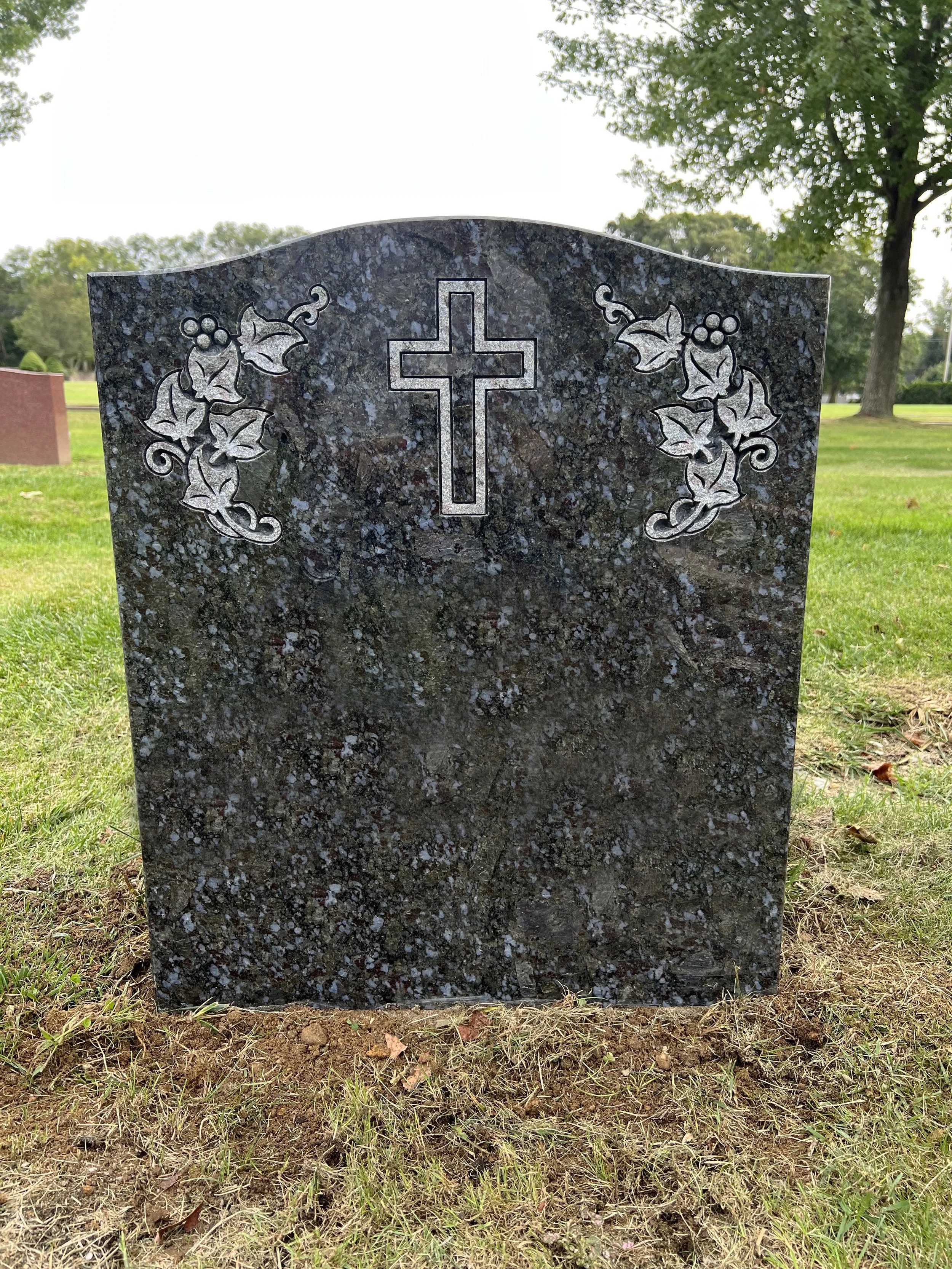 A black granite headstone with a Christian cross and floral carvings, set in a grassy cemetery with trees in the background.