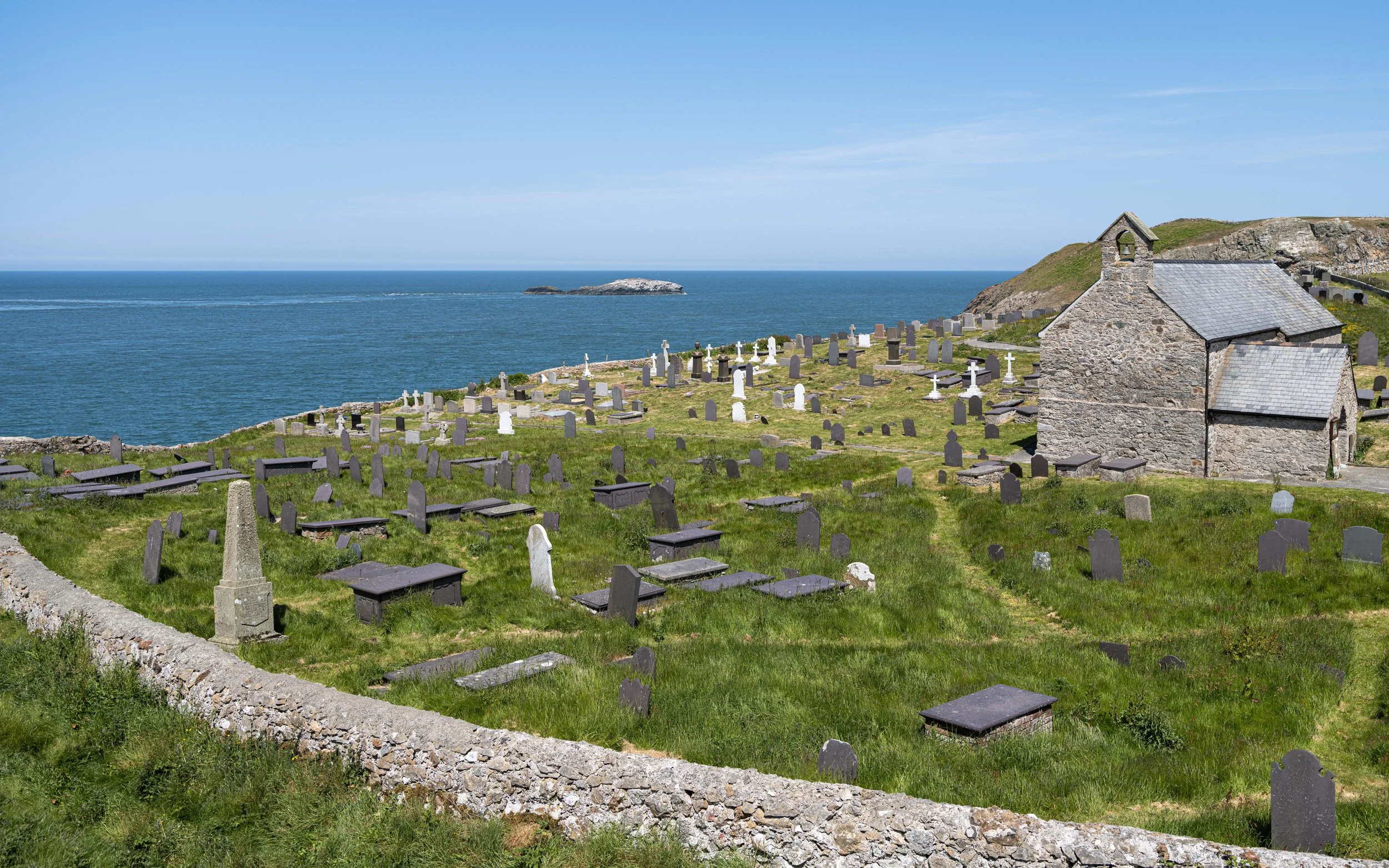 Grassy cemetery on a hill overlooking the ocean, with numerous gray and white tombstones and a small stone chapel, under a blue sky.