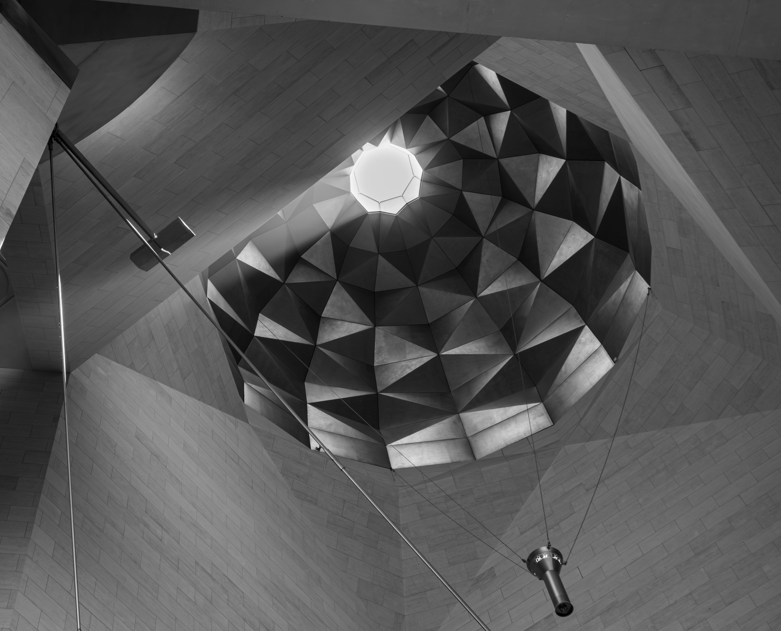 Interior view of a modern architectural ceiling with a geometric design, featuring a skylight at the center and a hanging security camera.