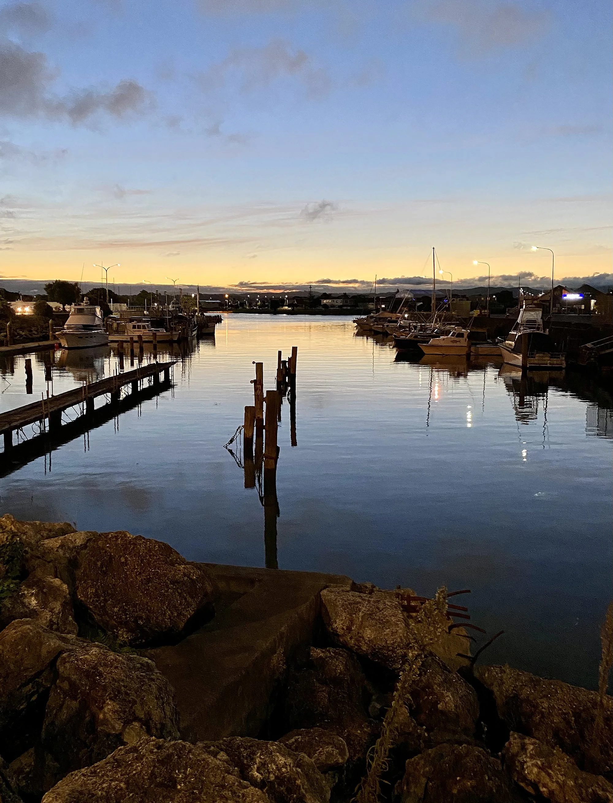 A serene marina at dusk with calm water, boats docked along both sides, a rocky shoreline in the foreground, and the sky transitioning from sunset to night with a few clouds overhead.