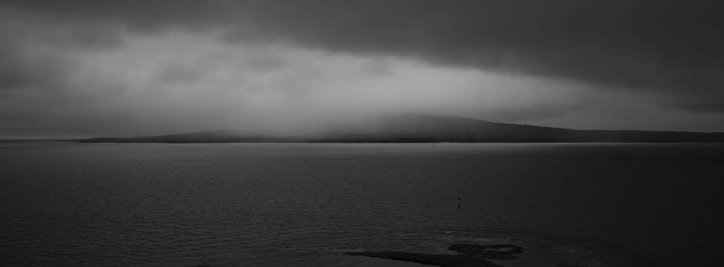 A black and white photograph of a calm body of water with a distant landmass under a cloudy sky.