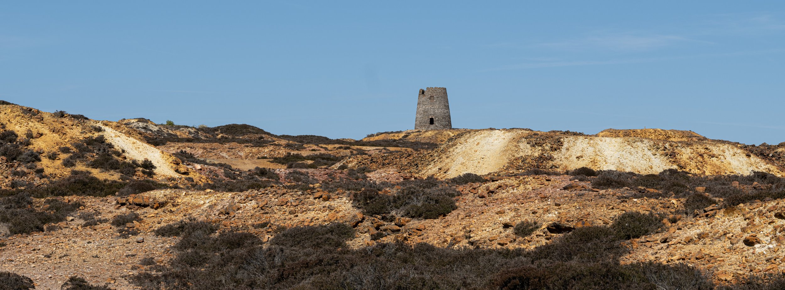 A tower on a hill in a desert landscape with rocky terrain and sparse vegetation, under a blue sky.