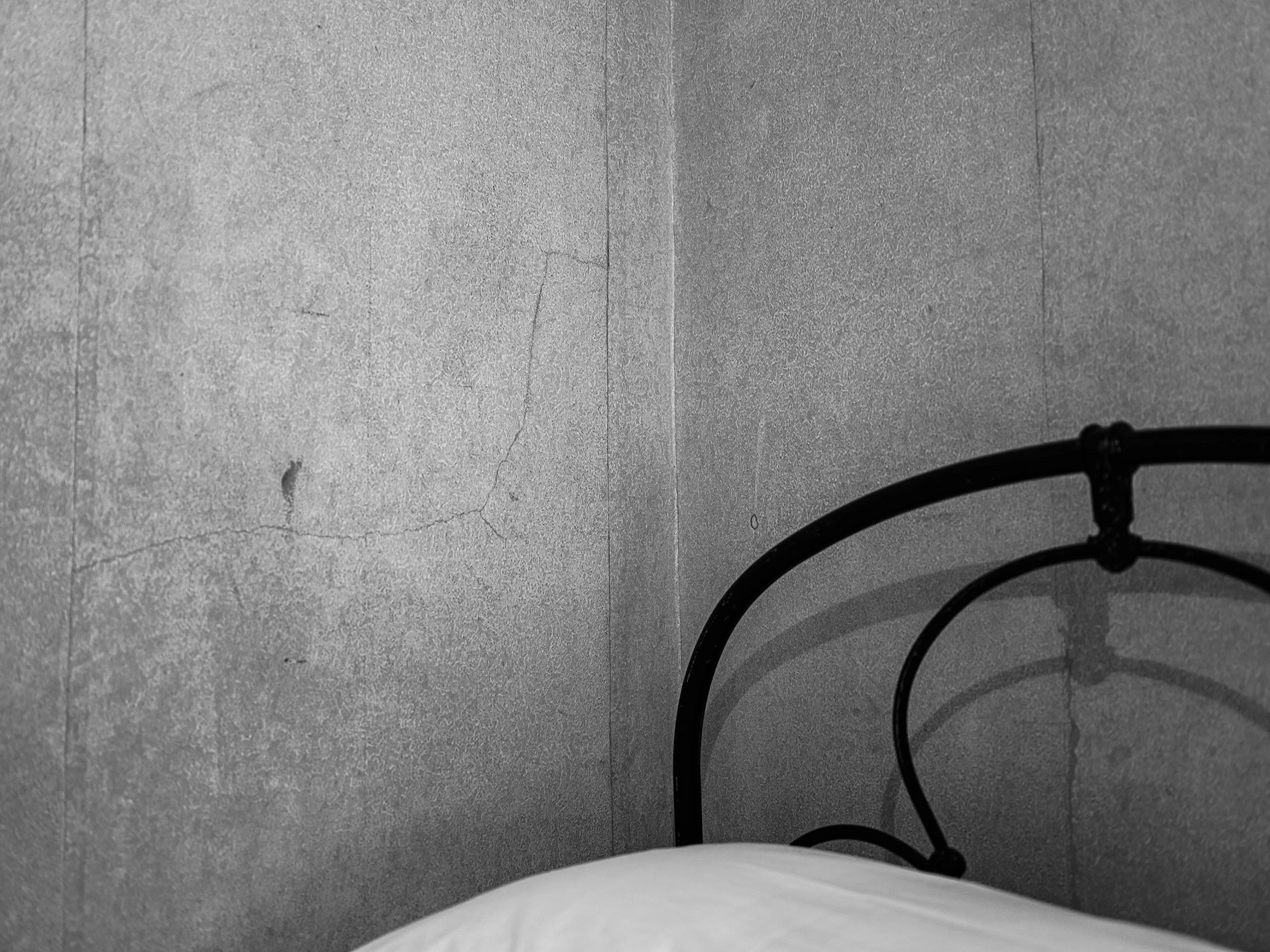 A black metal bed frame with a curved headboard is shown in the corner of a room with gray cracked concrete walls.