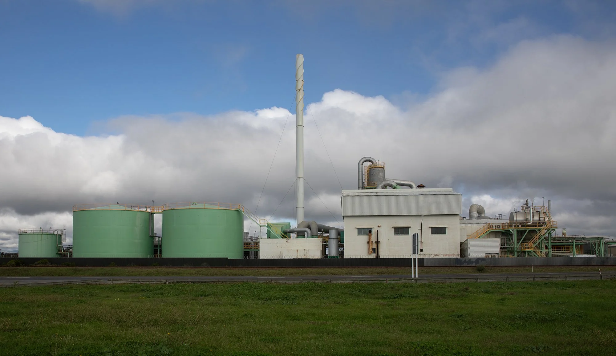 Industrial facility with large green storage tanks, a white building, and various pipes and structures under a cloudy sky.