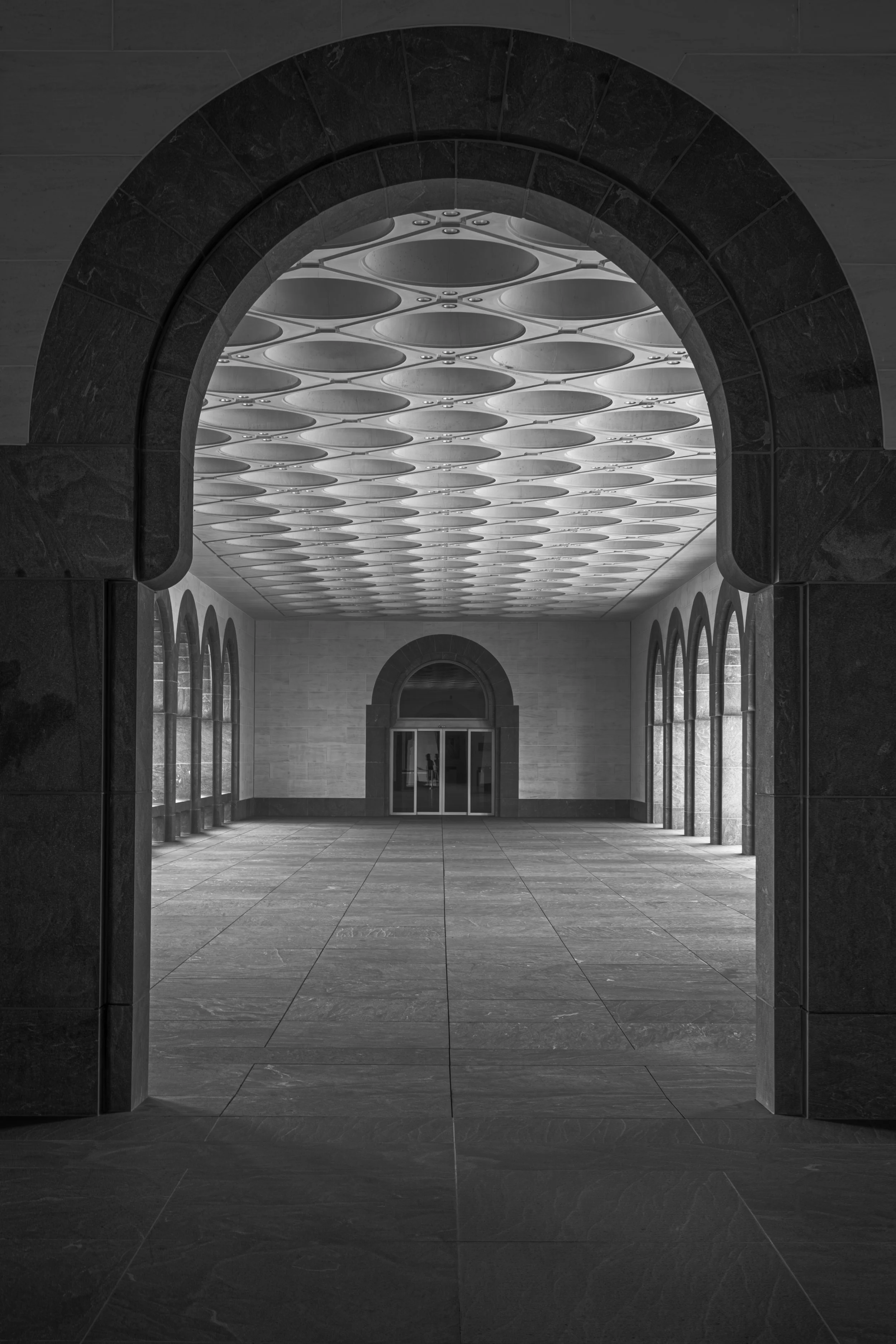 Interior view of a building with arched doorways, large windows on each side, a tiled floor, and a ceiling with circular patterned panels, in black and white.