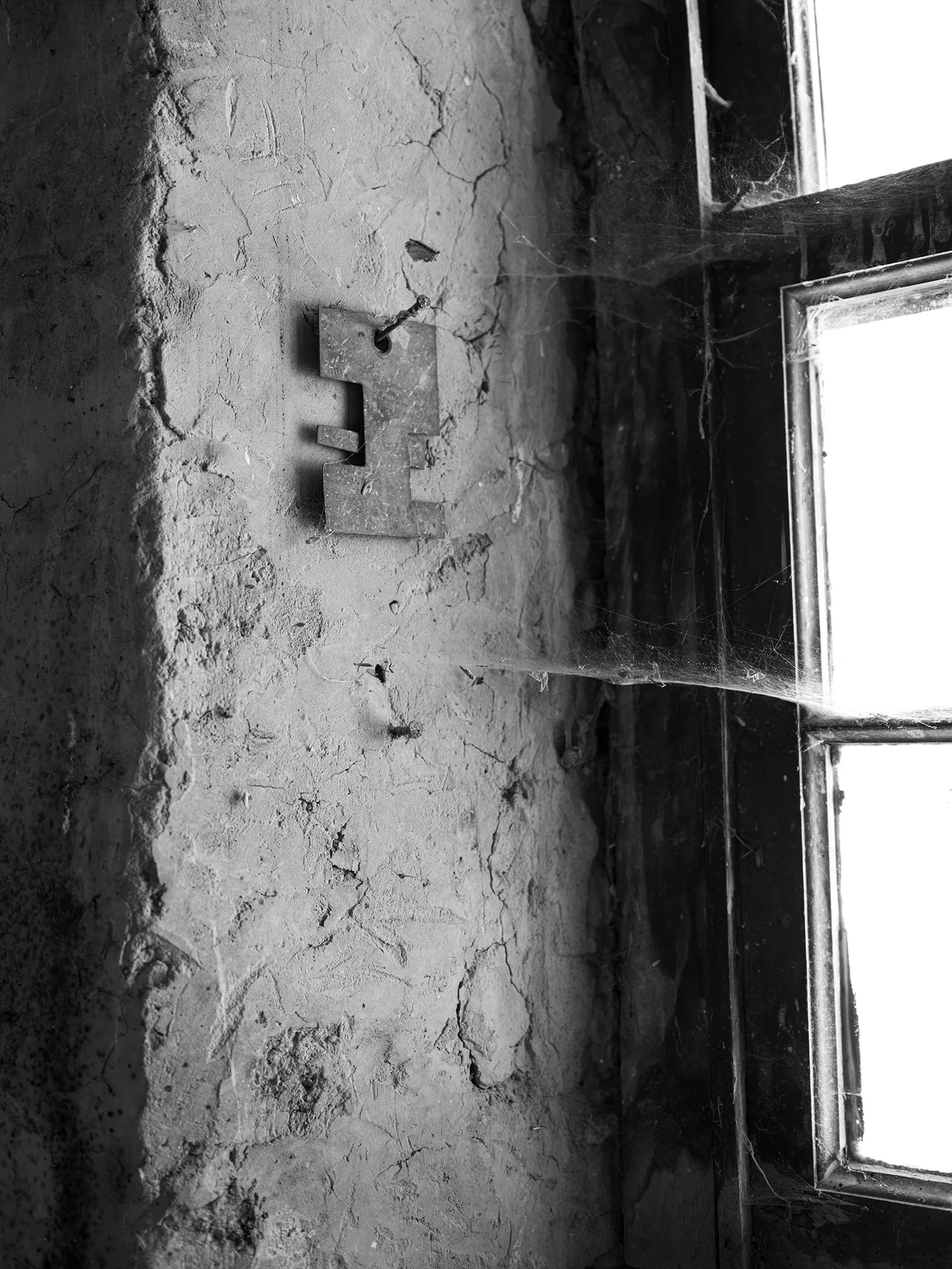 A black and white photo of a cracked, dusty window sill with spider webs and a metal letter 'E' attached to the wall near the window in an abandoned building.