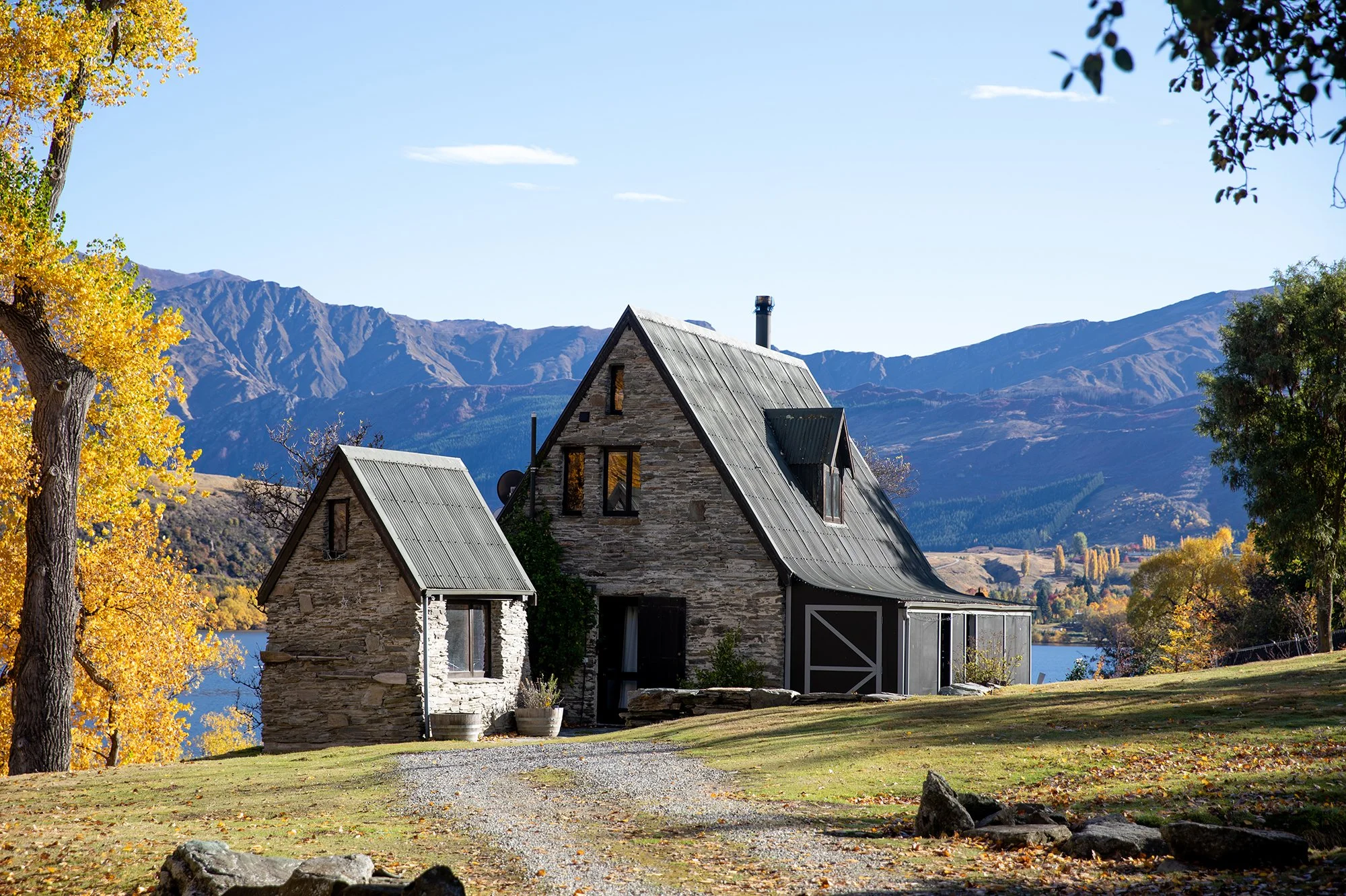 Stone cabin and smaller stone structure with metal roofs, surrounded by autumn trees, a lake, and mountain range in the background.