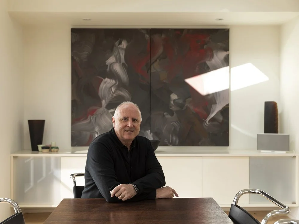 A man with gray hair sitting at a wooden table in a modern room, with abstract art and vases on a white cabinet behind him.