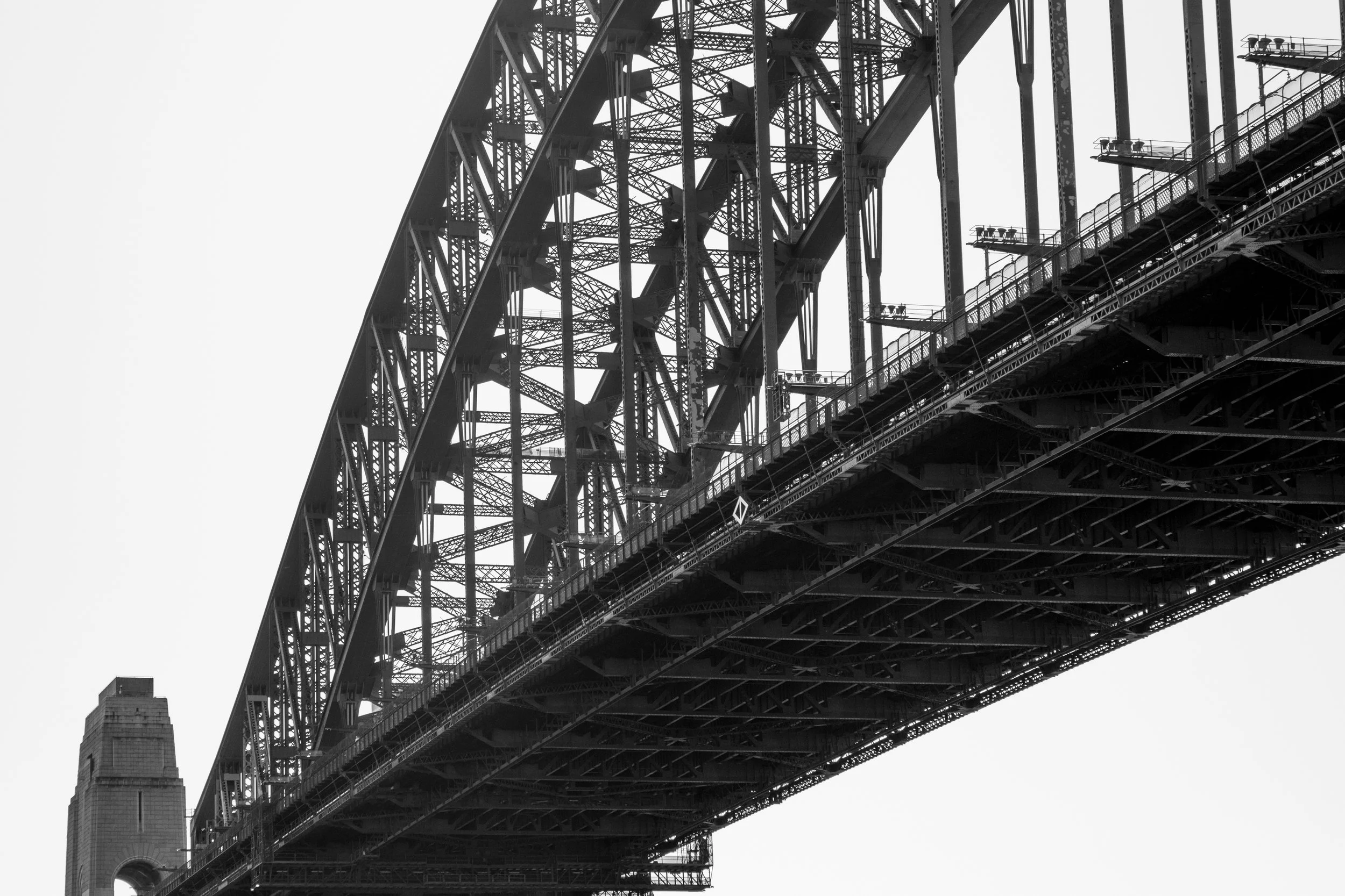 Black and white photo of the underside of a large bridge showing intricate steel framework and part of a building in the background.