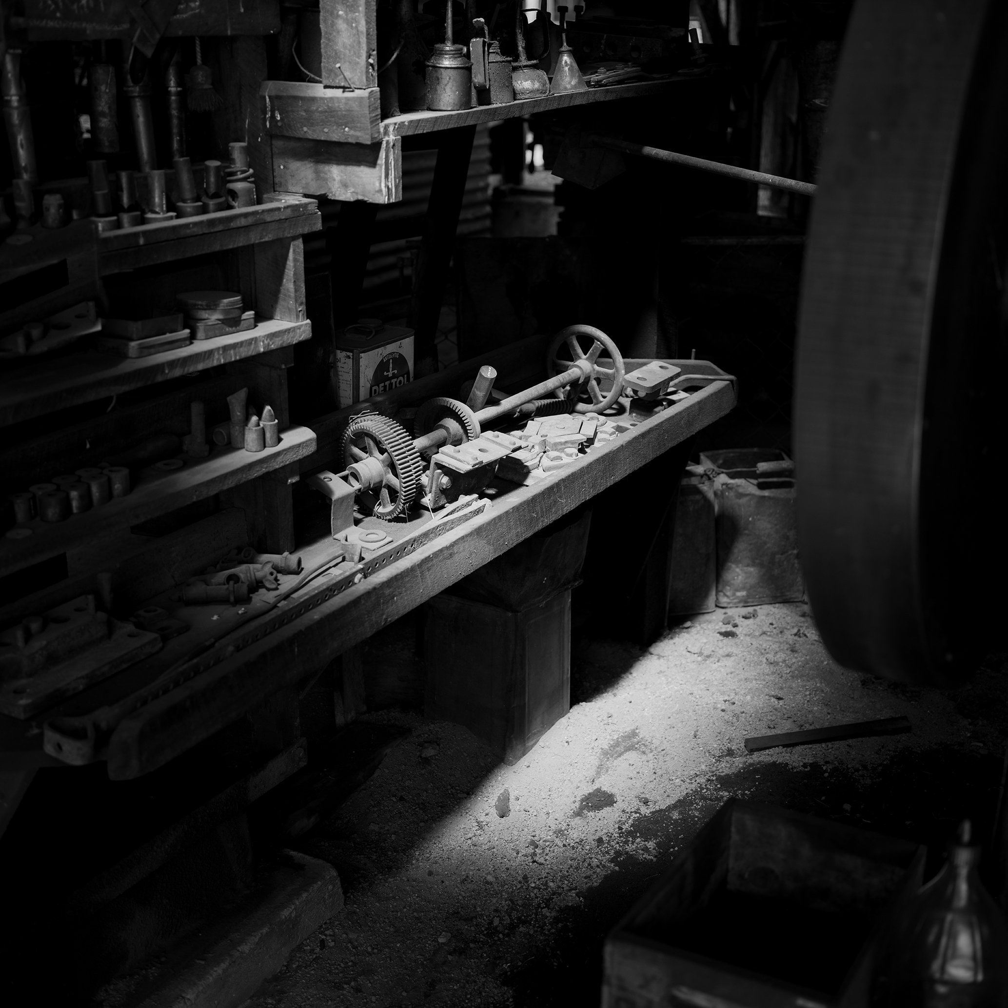 Black and white photo of a cluttered woodworking workshop with various tools and machine parts on a workbench, illuminated by a beam of light on the dusty floor.