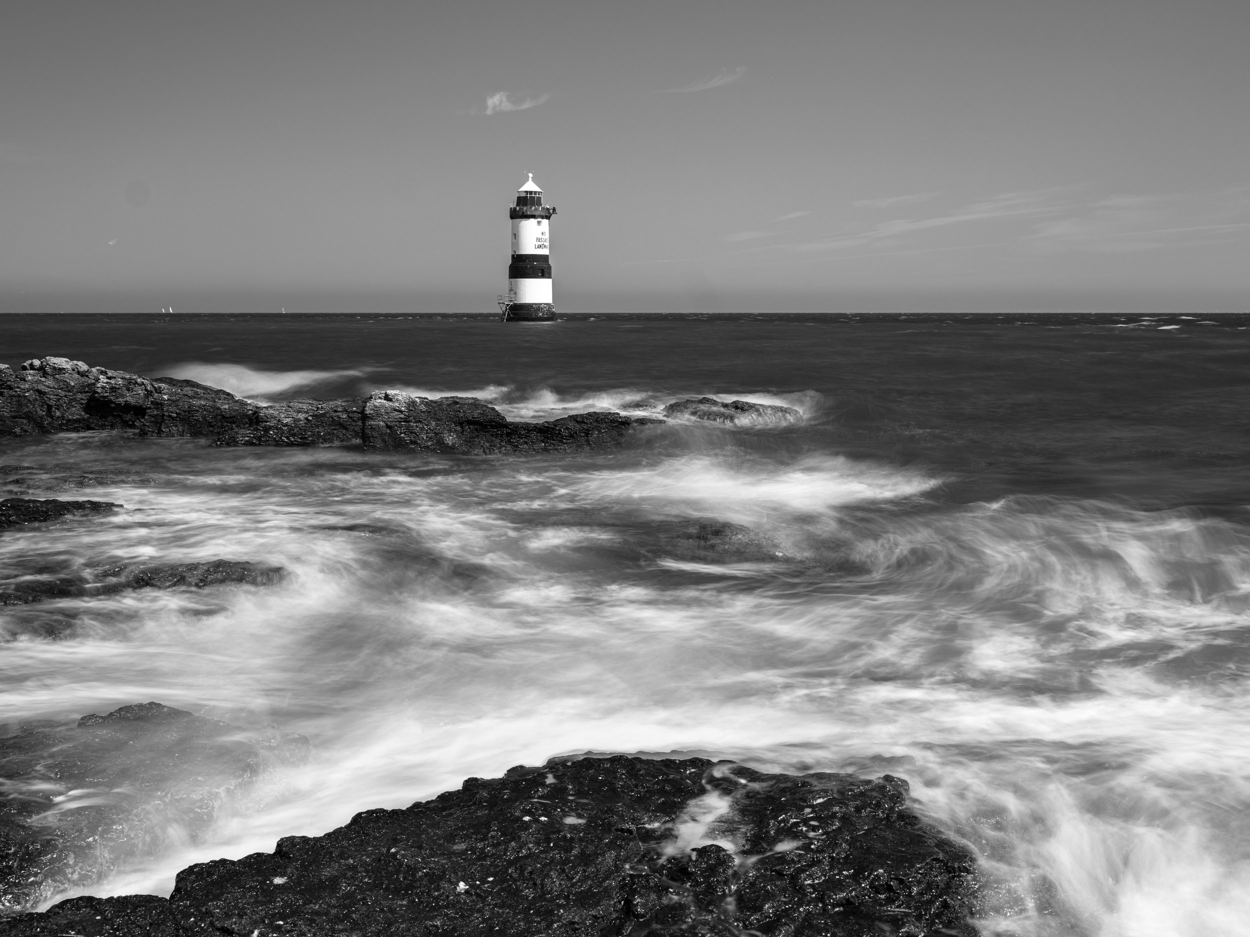 Black and white photo of a lighthouse in the distance over ocean waves crashing against rocks in the foreground.