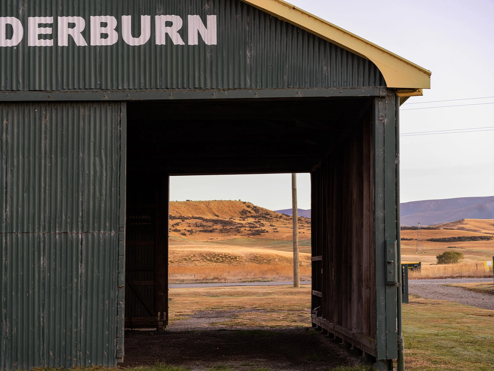 View through the open entrance of a green corrugated metal barn with distant rolling hills and a partly cloudy sky in the background.