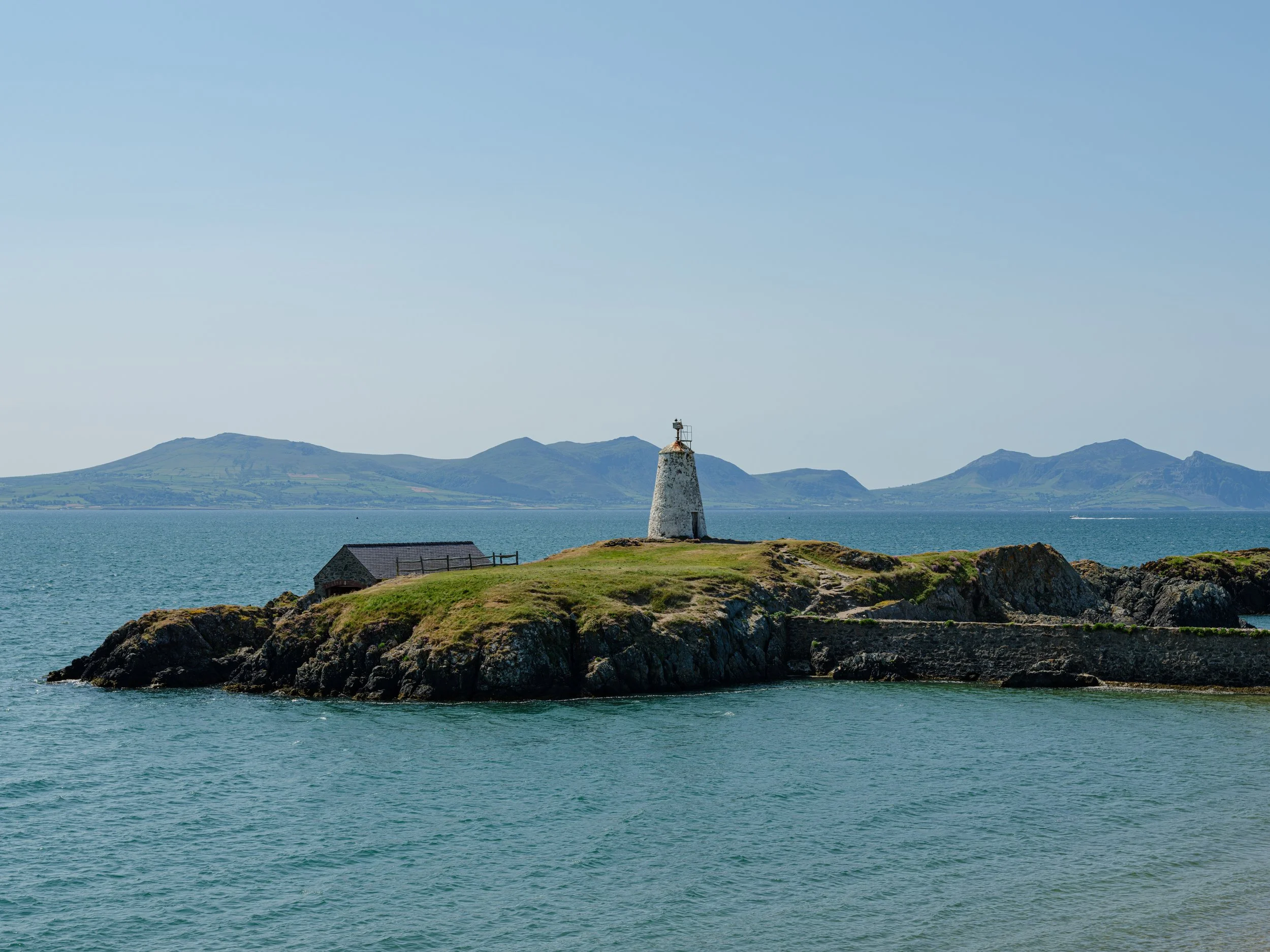 A small island with a lighthouse and a house, surrounded by water, with distant hills or mountains in the background.