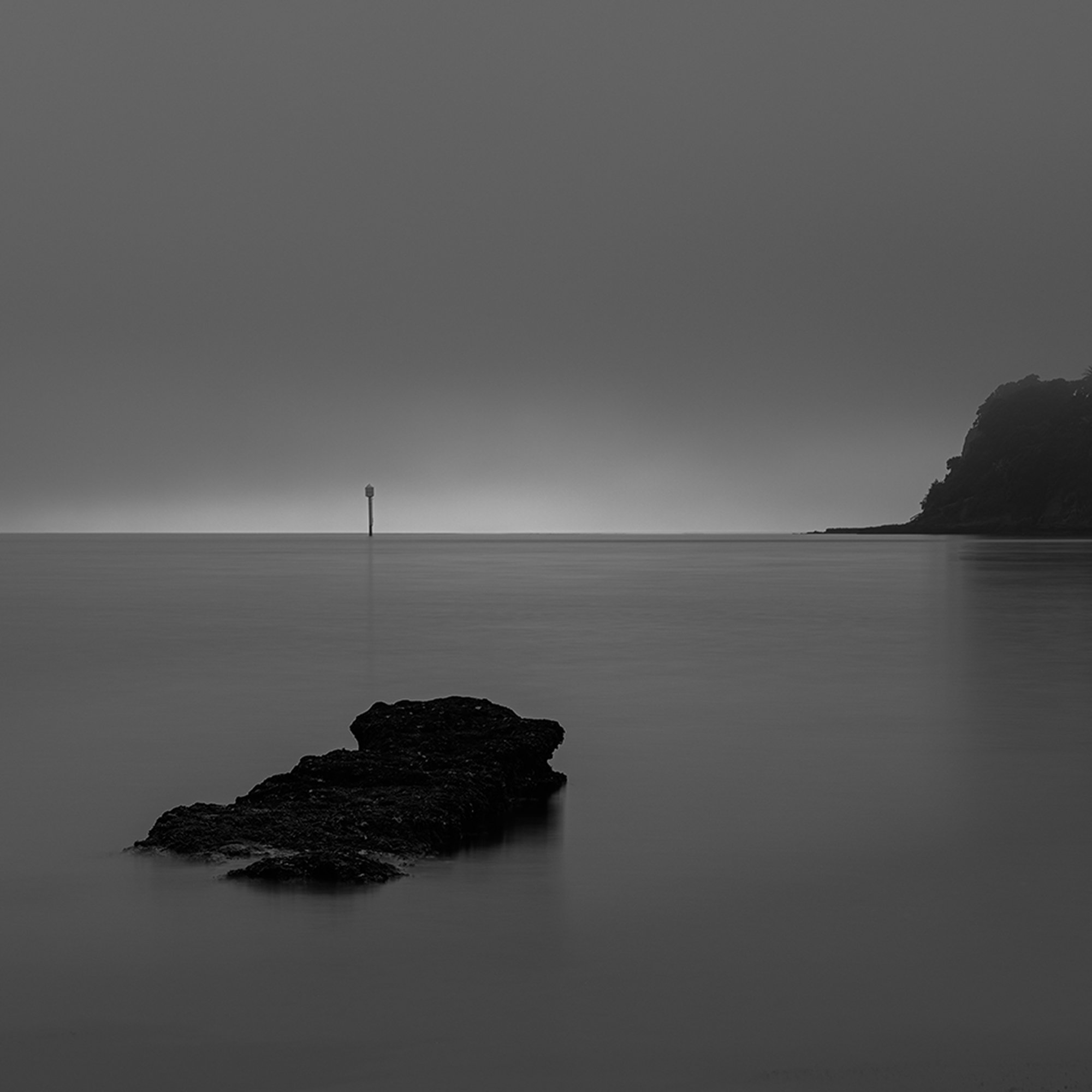 Black and white photo of a calm ocean with a rock in the foreground, a distant navigation marker in the middle, and a rocky headland on the right.