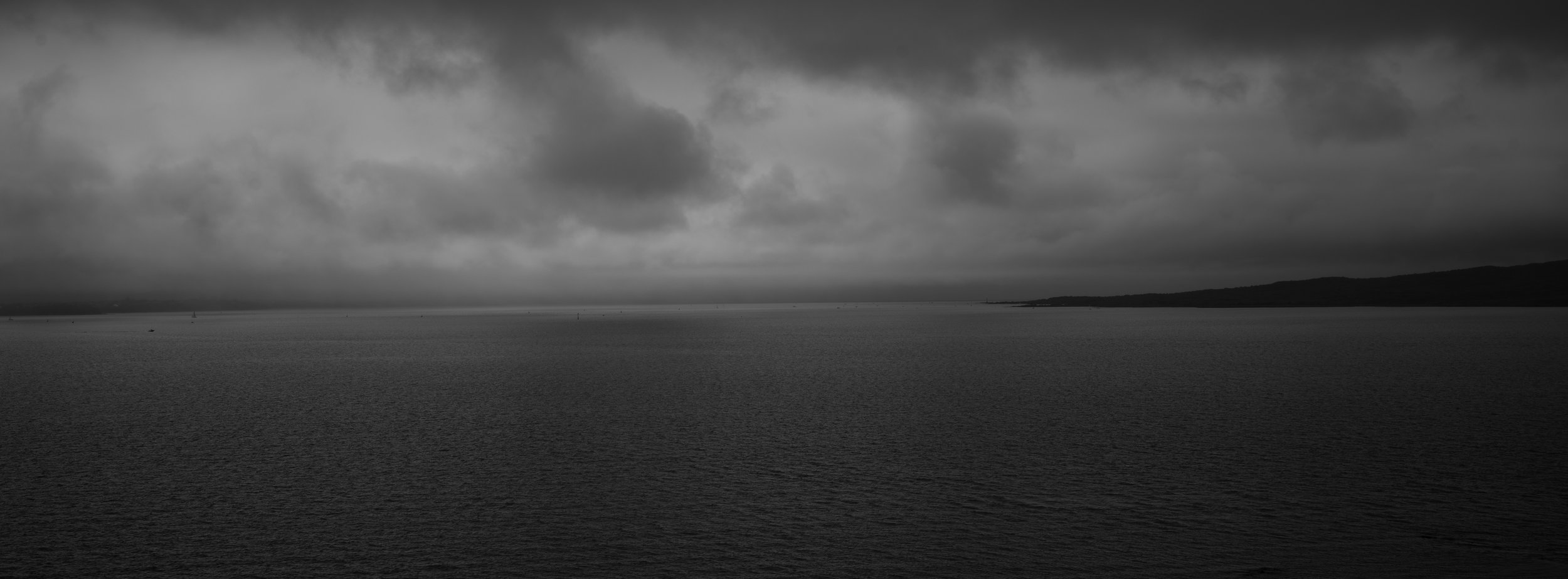 Black and white photo of a large body of water with a distant shoreline and hills under a cloudy sky.