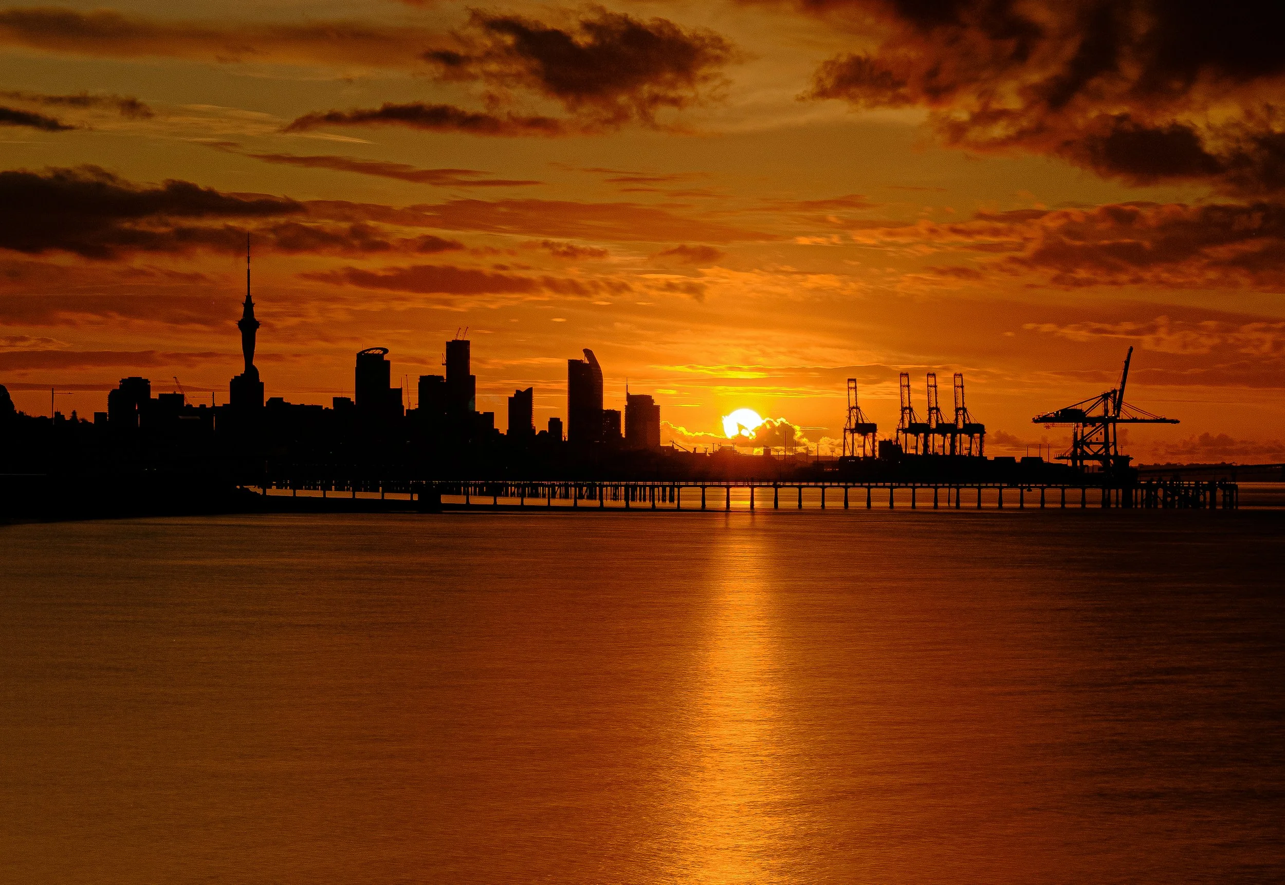 A silhouette of a city skyline with tall buildings and cranes at sunset, with the sun partially visible on the horizon, reflecting on calm water in the foreground, and a partly cloudy sky.