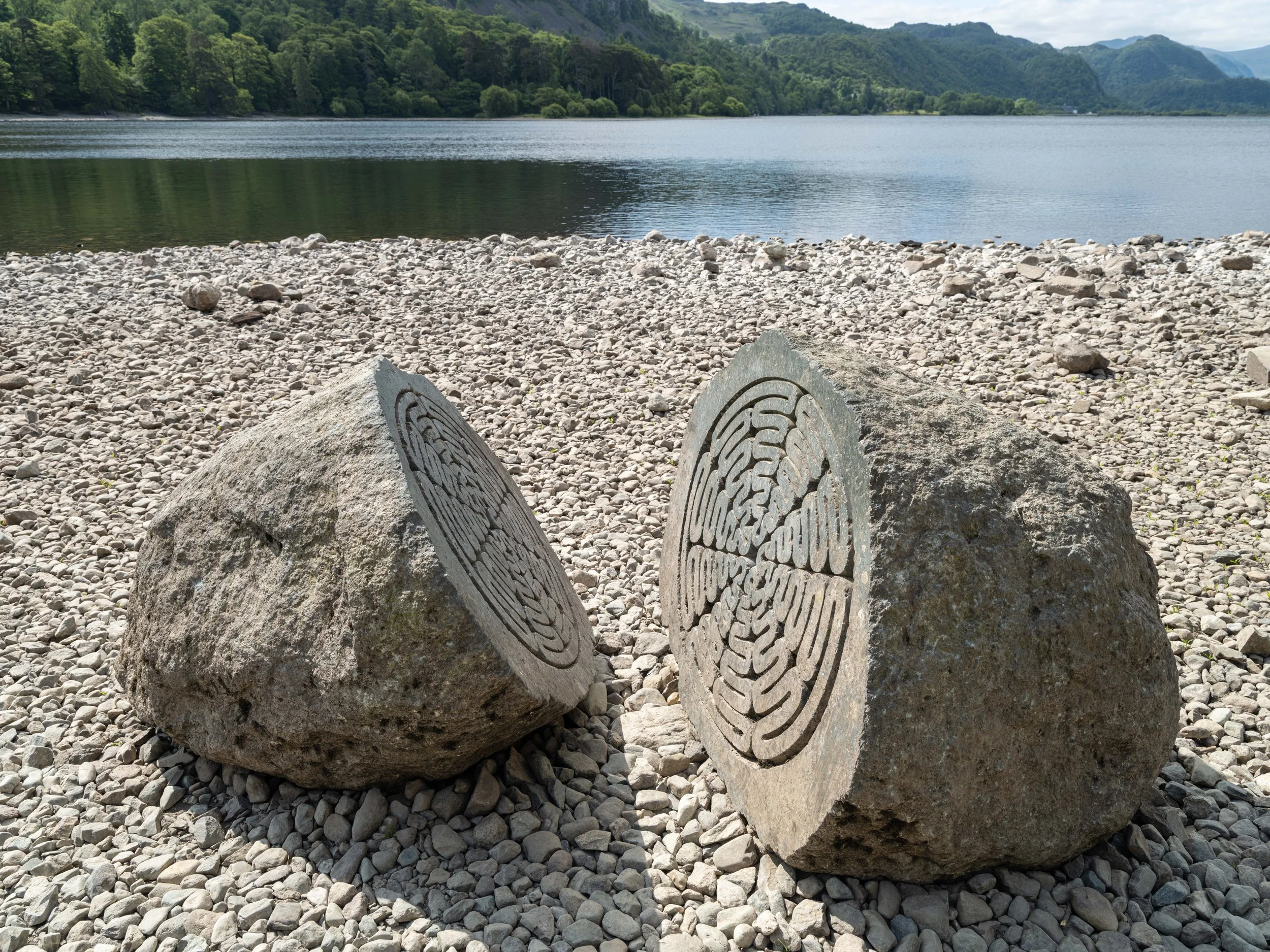 Two large rocks with carved petroglyphs sit on a pebbled shoreline near a body of water, with green hills and mountains in the background.
