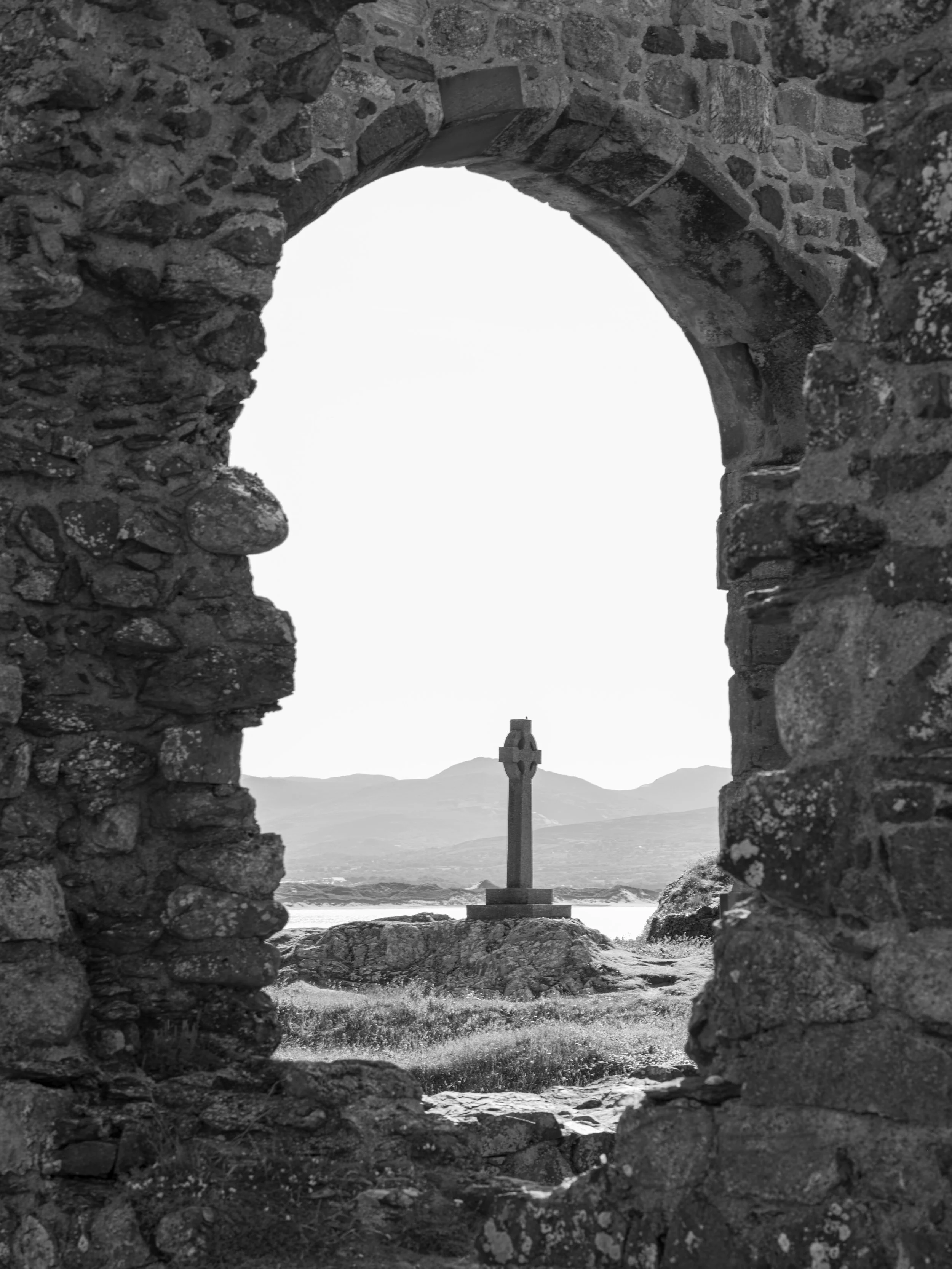 A view through an arched stone window framing a Celtic cross monument on rocky ground with mountains and water in the background.