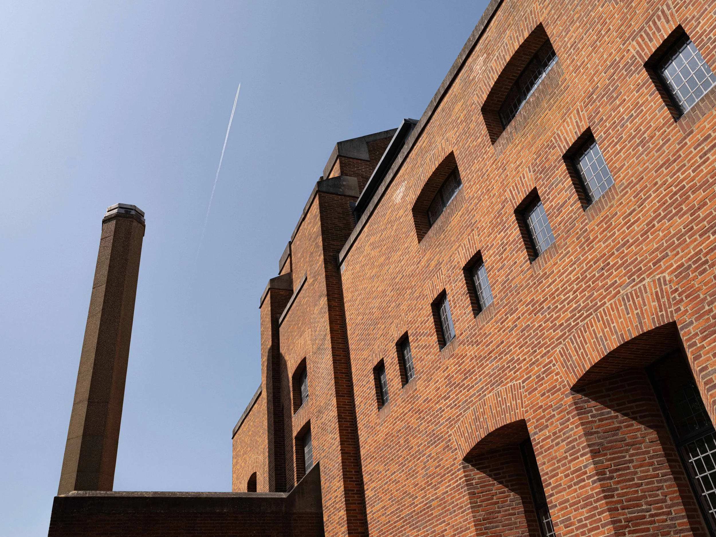 A large brick building with multiple windows and a tall brick chimney under a clear blue sky with a jet contrail.