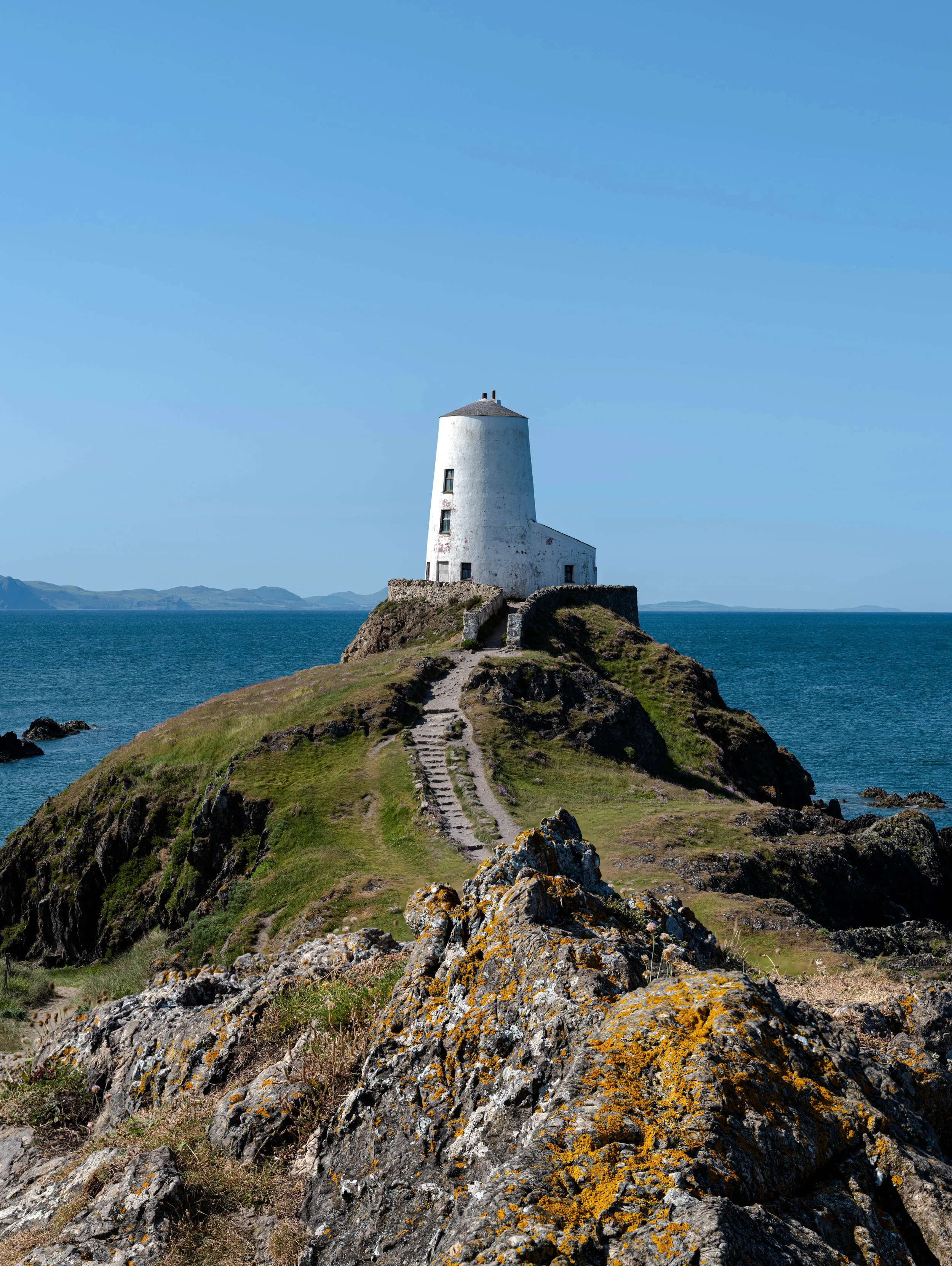 A white lighthouse on a rocky hill overlooking the ocean, with a clear blue sky and distant land on the horizon.