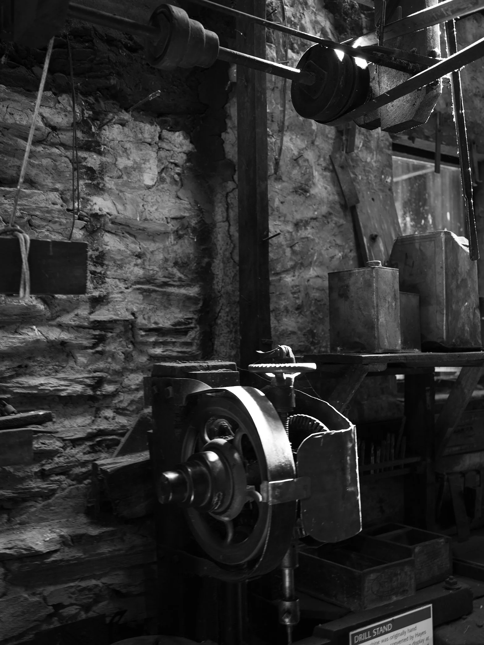 Vintage black and white image of a workshop with an old drill press, wooden shelves, and stone wall in the background.