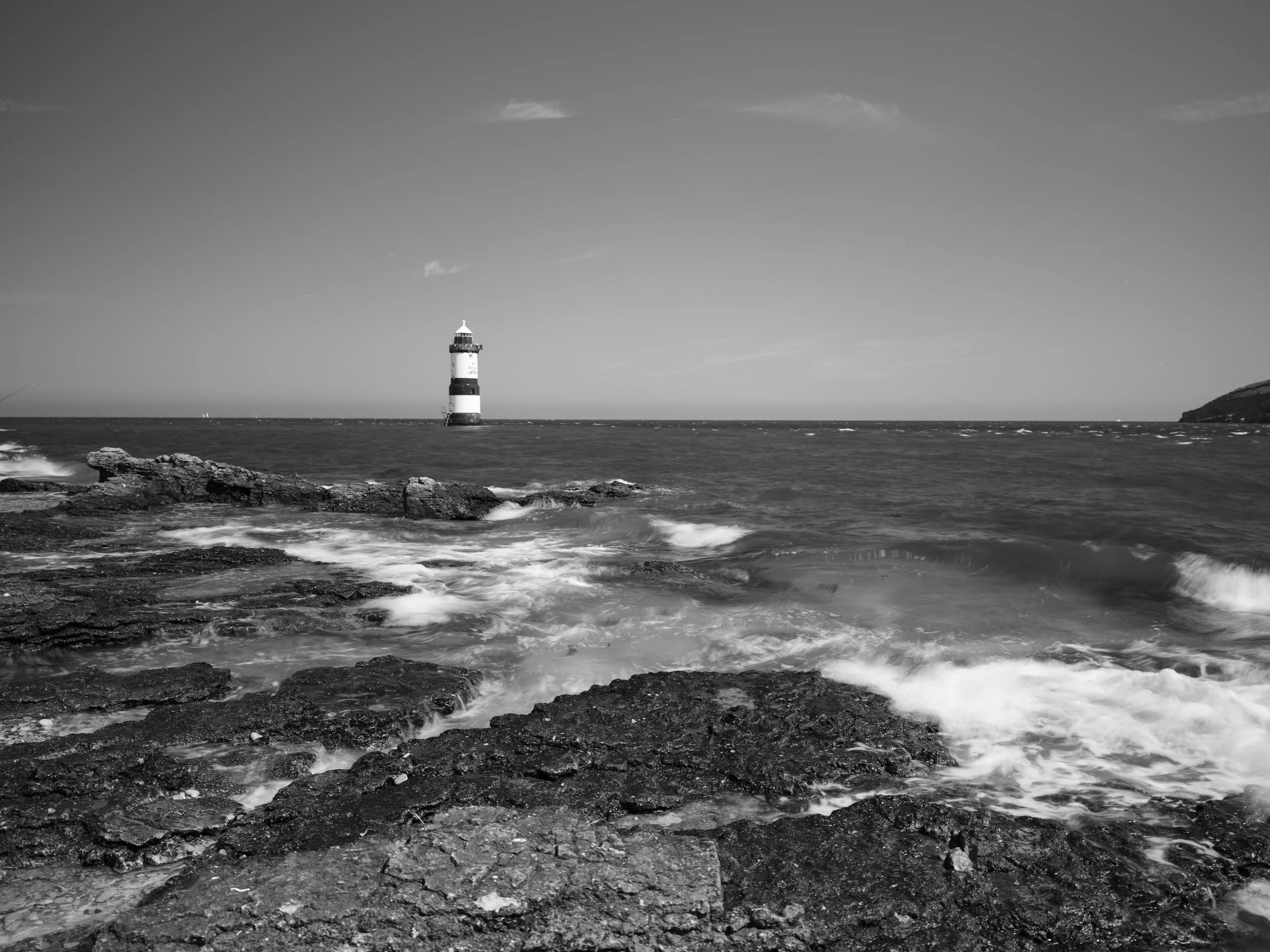 Black and white photo of a lighthouse on the ocean, with rocky shoreline in foreground and waves crashing.