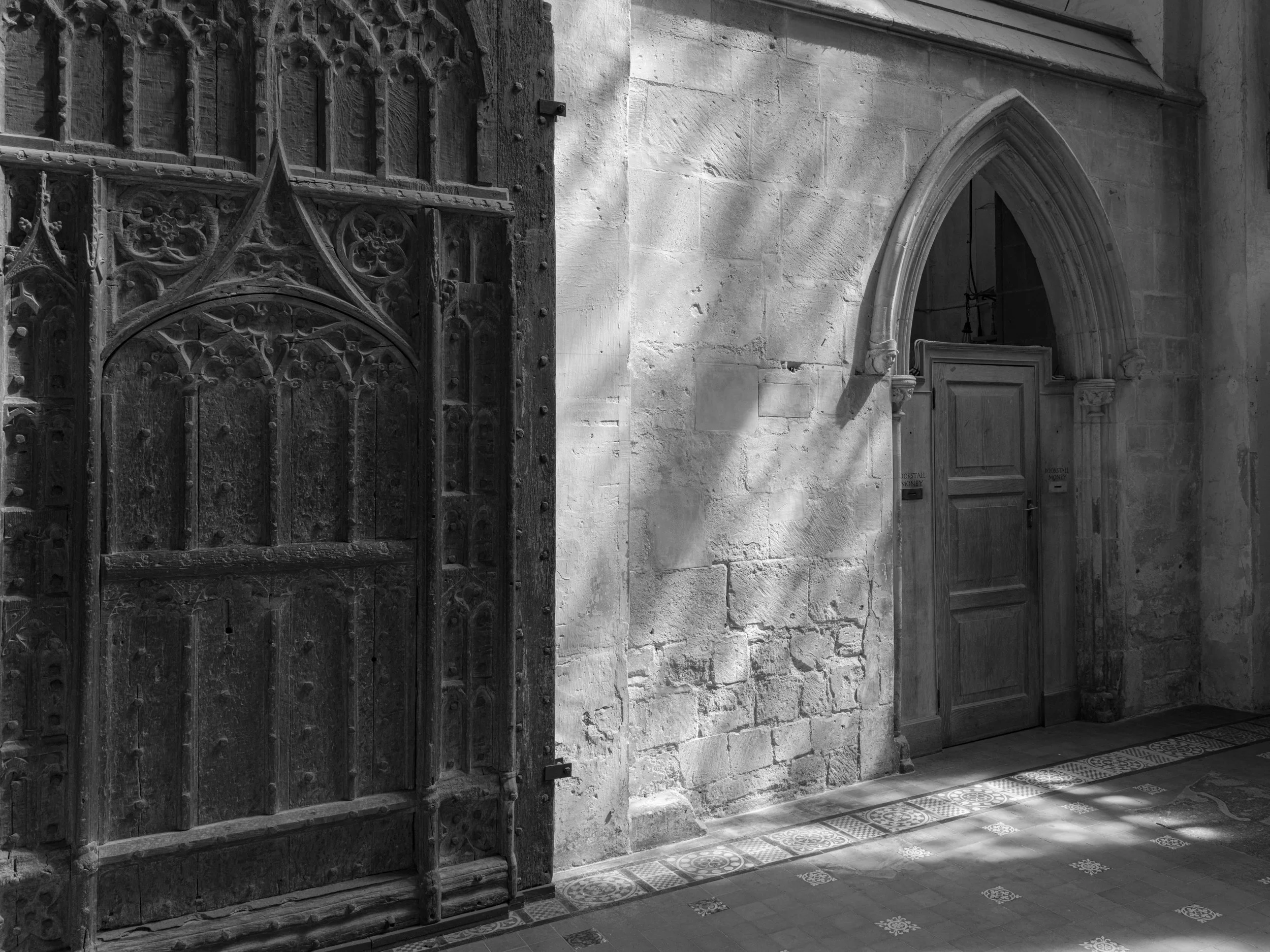 Black and white image of an ornate wooden door with Gothic architecture, a pointed arch doorway, and stone wall with intricate carvings, sunlight casting shadows on the floor.