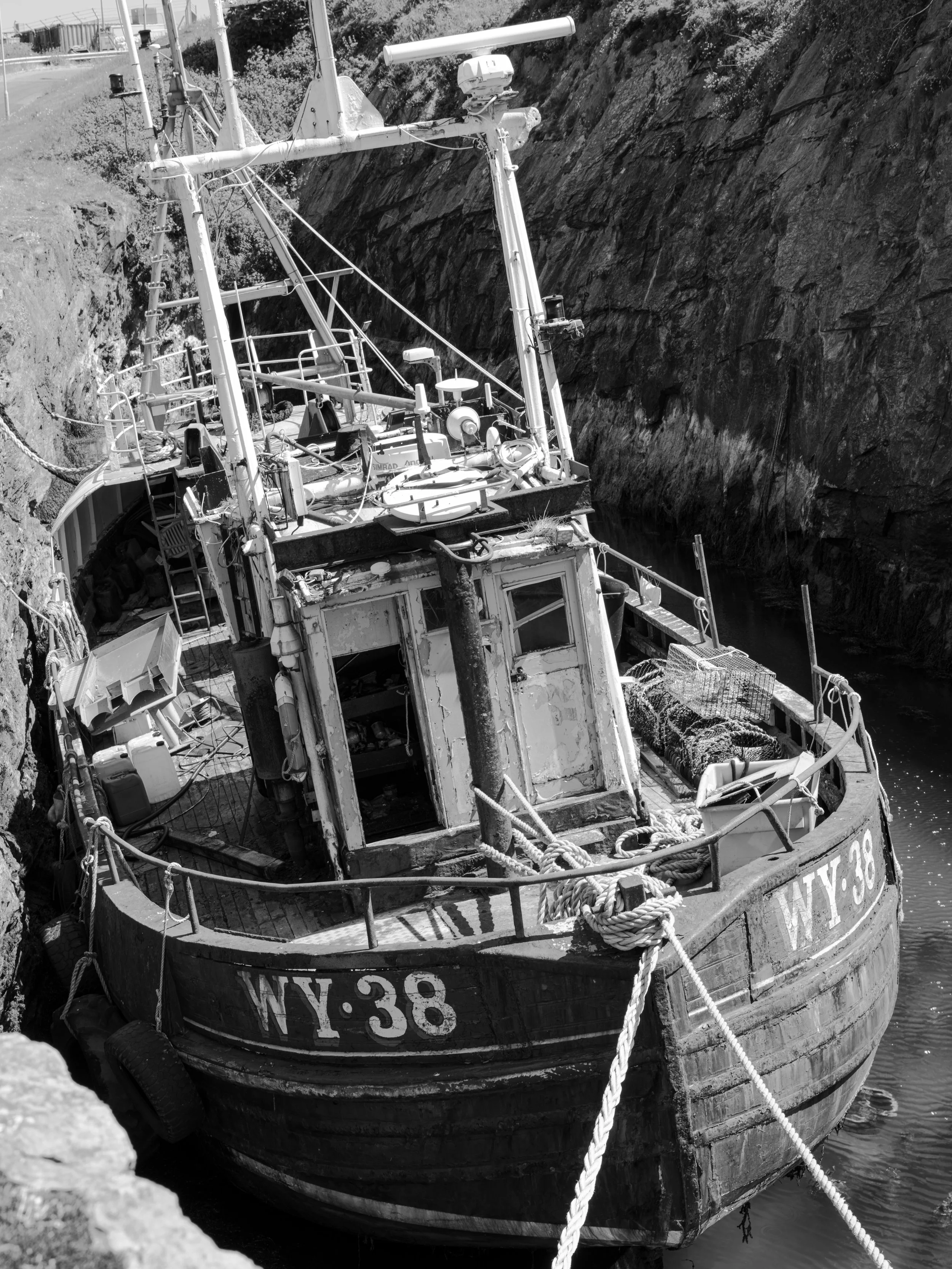 An old, weathered boat labeled WY-38, moored in a narrow waterway between rocky cliffs, with various equipment and ropes on deck.