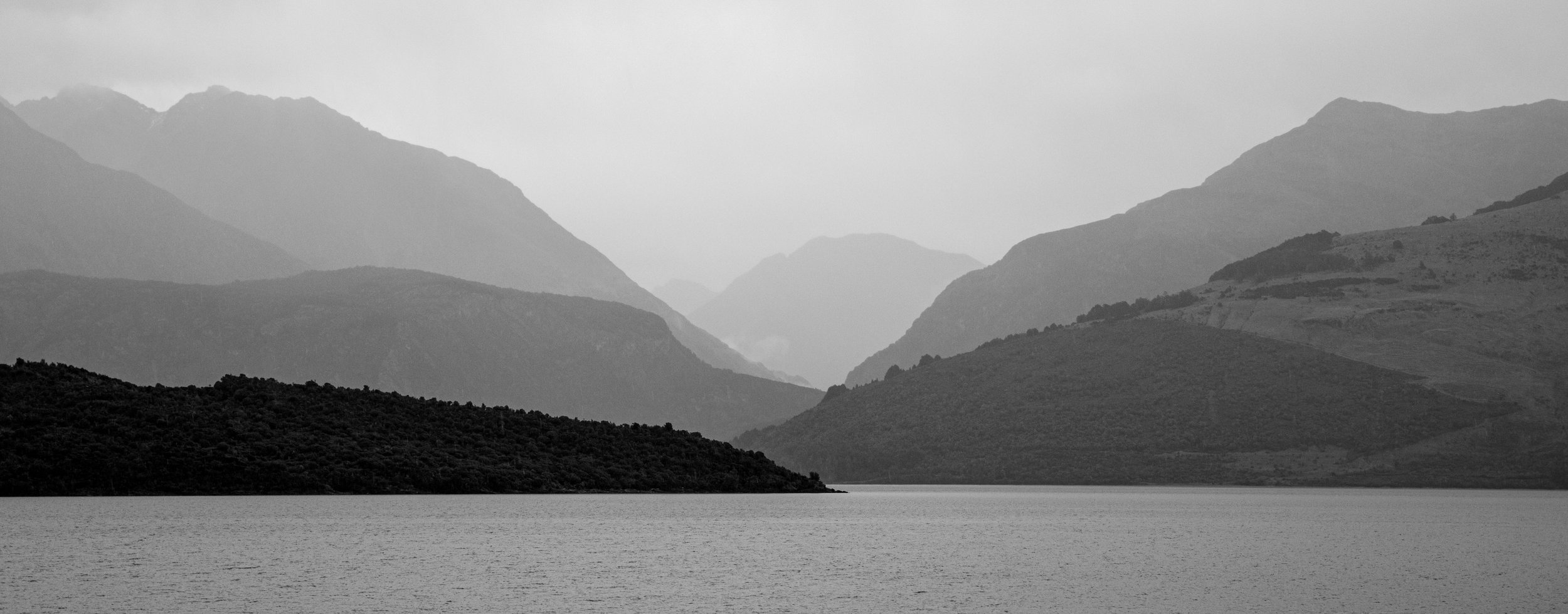 Black and white photo of a large lake with surrounding mountains in the background, mist or fog partially obscuring the peaks.
