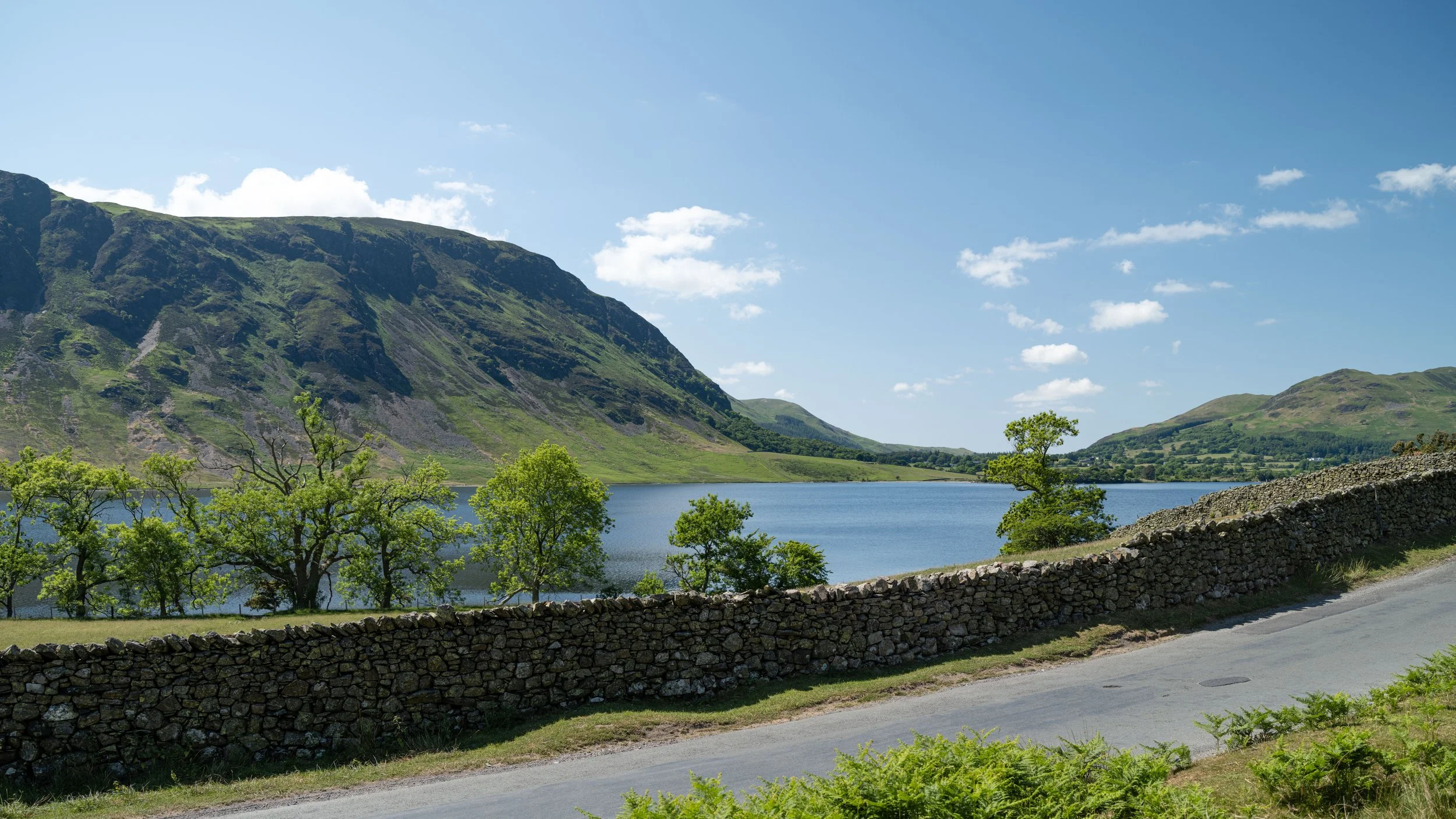 A scenic landscape with a lake, green trees, and rolling hills under a partly cloudy blue sky.