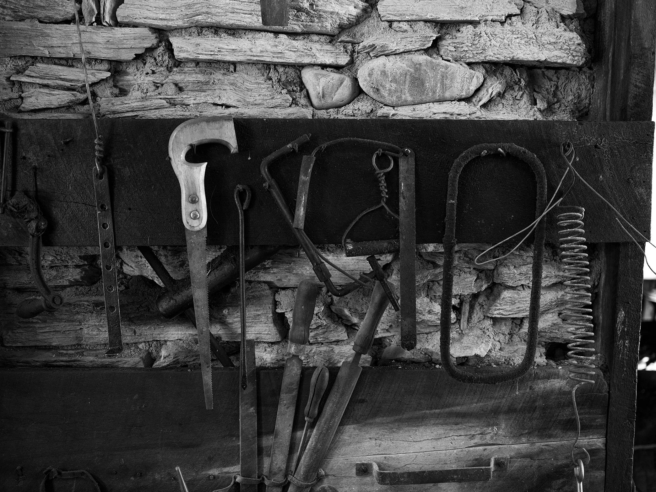 Tools hanging on a wooden board against a stone wall, including saws, a hammer, a hook, and a spring.