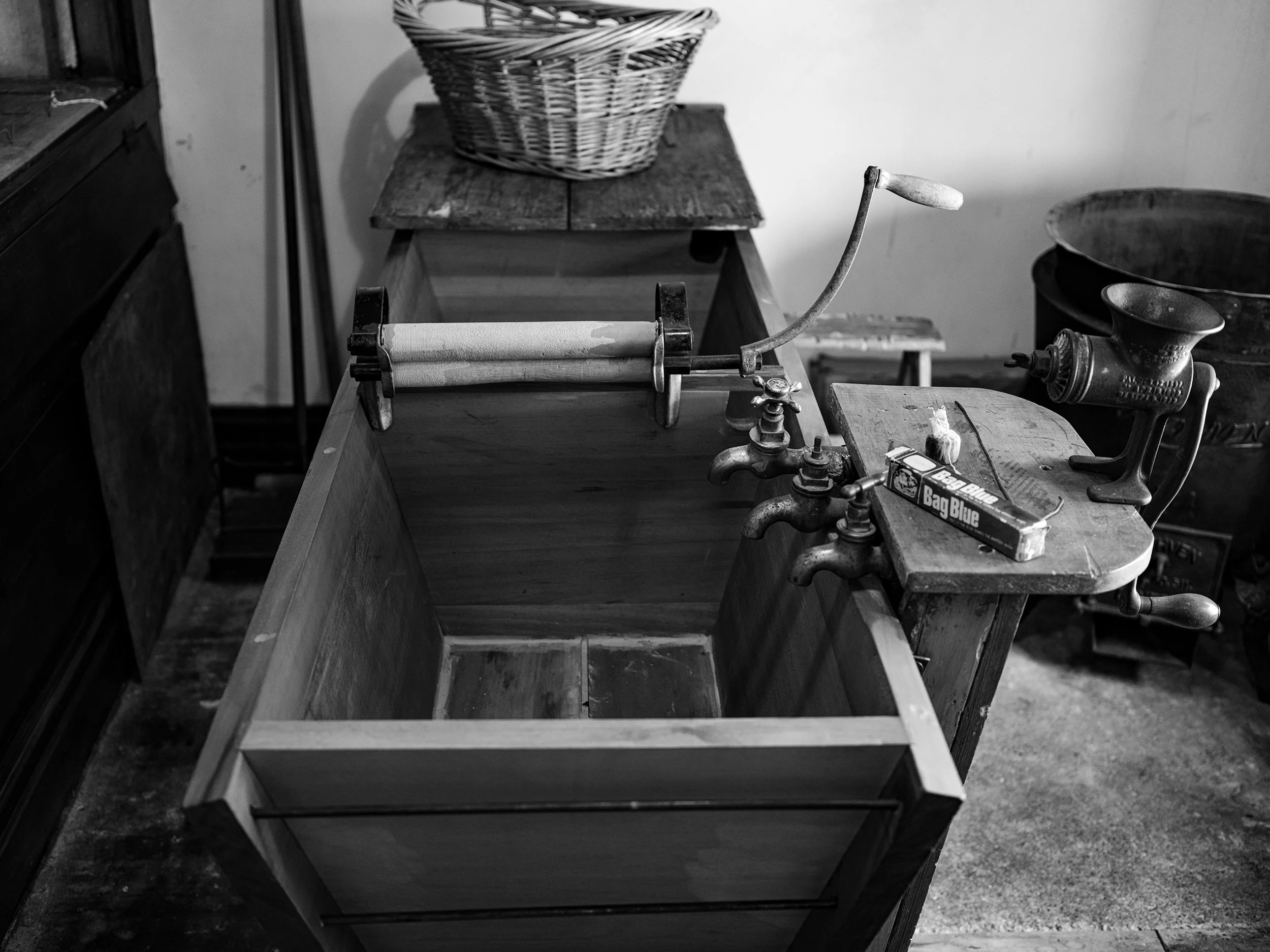 Old wooden washbasin with vintage faucet and hand-crank wringer, surrounded by antique kitchen items in black and white.